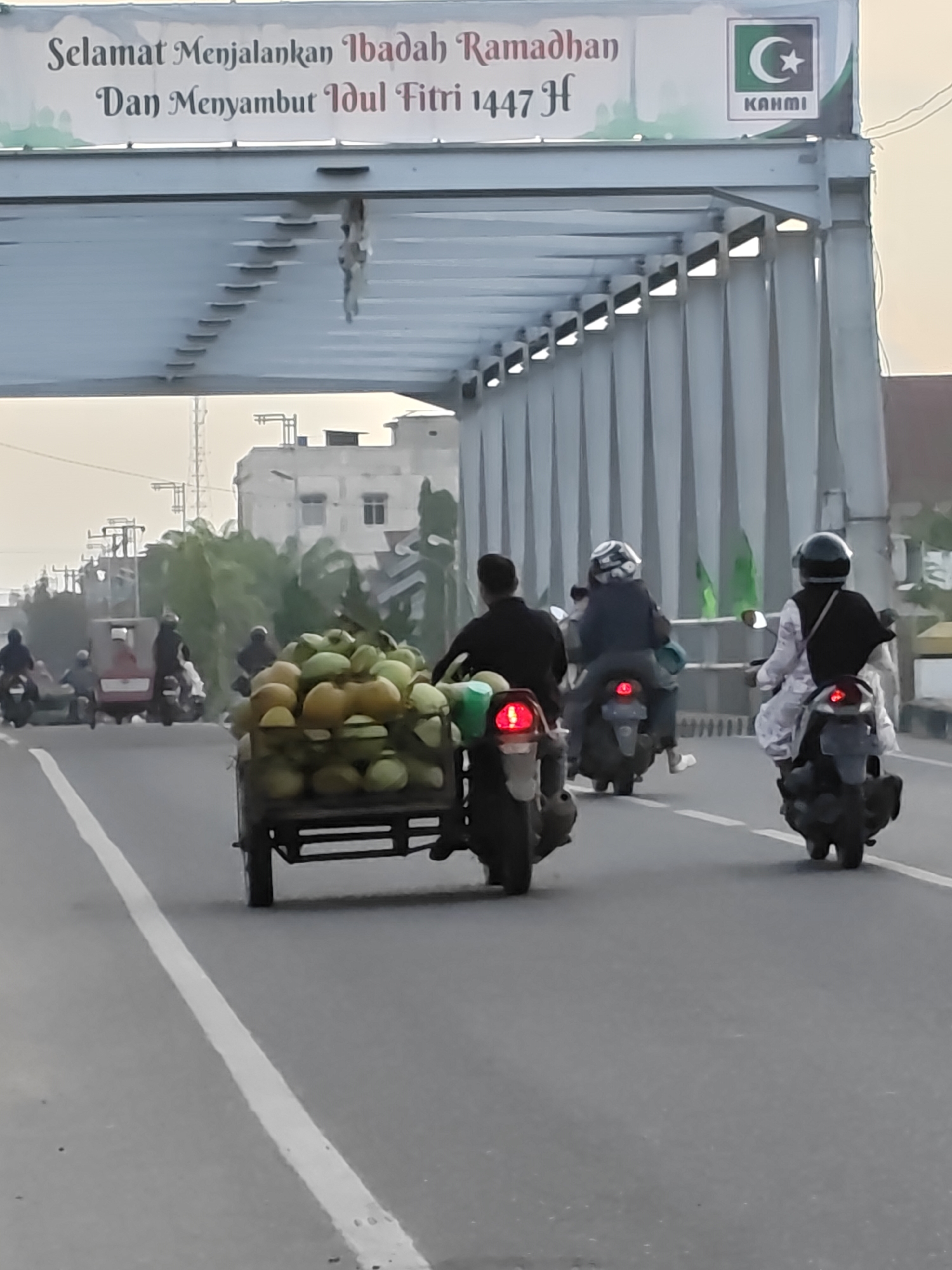 The image shows a street scene in Lhokseumawe, Indonesia. A motorized tricycle laden with green coconuts is the focal point, moving along a gray asphalt road. Behind it, two motorcycles are also traveling in the same direction. Additional traffic consisting of motorbikes is visible in the distance. The sky is overcast, casting a soft light on the scene. Overhead, a bridge with white painted steel beams is prominently displayed with a banner reading "Selamat Menjalankan Ibadah Ramadhan Dan Menyambut Idul Fitri 1447 H," indicating the celebration of Ramadan and Eid al-Fitr in the Islamic calendar. The banner also includes a symbol of a crescent moon and star. Buildings and trees can be seen in the background.