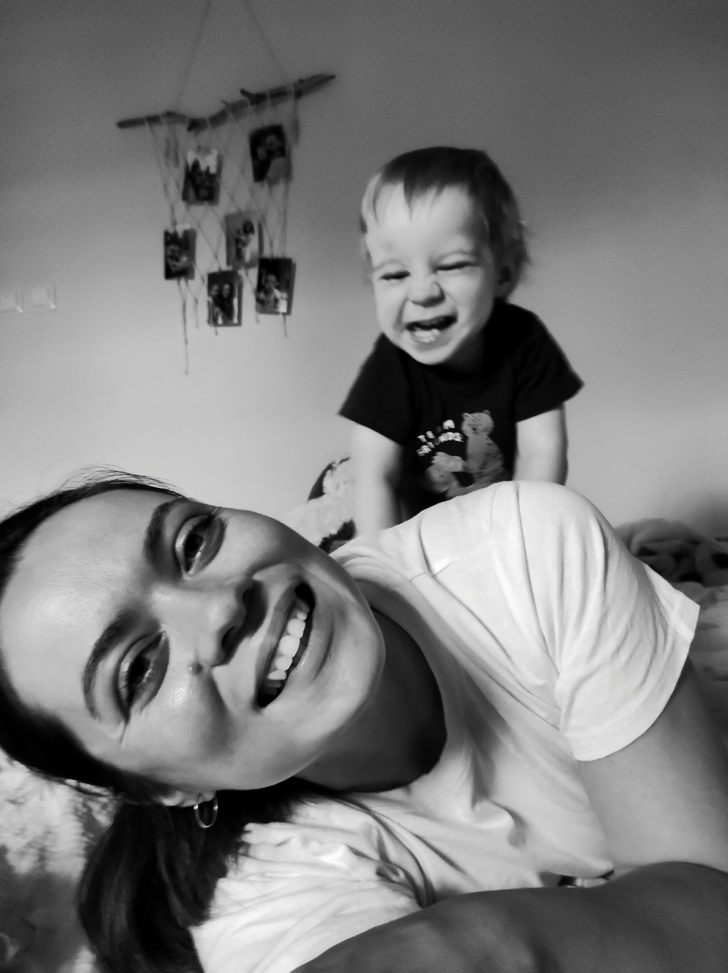 A black and white photograph captures a candid and joyful moment between a young woman and a toddler in an indoor setting, likely a home in Fokino, Russia. The composition places the woman prominently in the foreground, smiling broadly and looking directly at the camera. Her head is tilted, showcasing a wide, toothy grin and bright eyes, conveying happiness and affection. She has dark hair and wears a light-colored t-shirt and a small hoop earring.
Behind her, a young child with light hair is visible, laughing with eyes squinted and mouth open wide, reflecting intense delight. The child wears a dark t-shirt with a faint, illegible design on the front, possibly a cartoon bear or text.
The background features a simple wall adorned with a rustic, homemade photo display. This decoration consists of a horizontal piece of wood or branch, from which several strings hang, each holding small, dark-framed photographs. The overall scene exudes a warm, playful, and intimate atmosphere. The monochrome palette emphasizes the emotional expressions of both subjects and the tender connection between them.