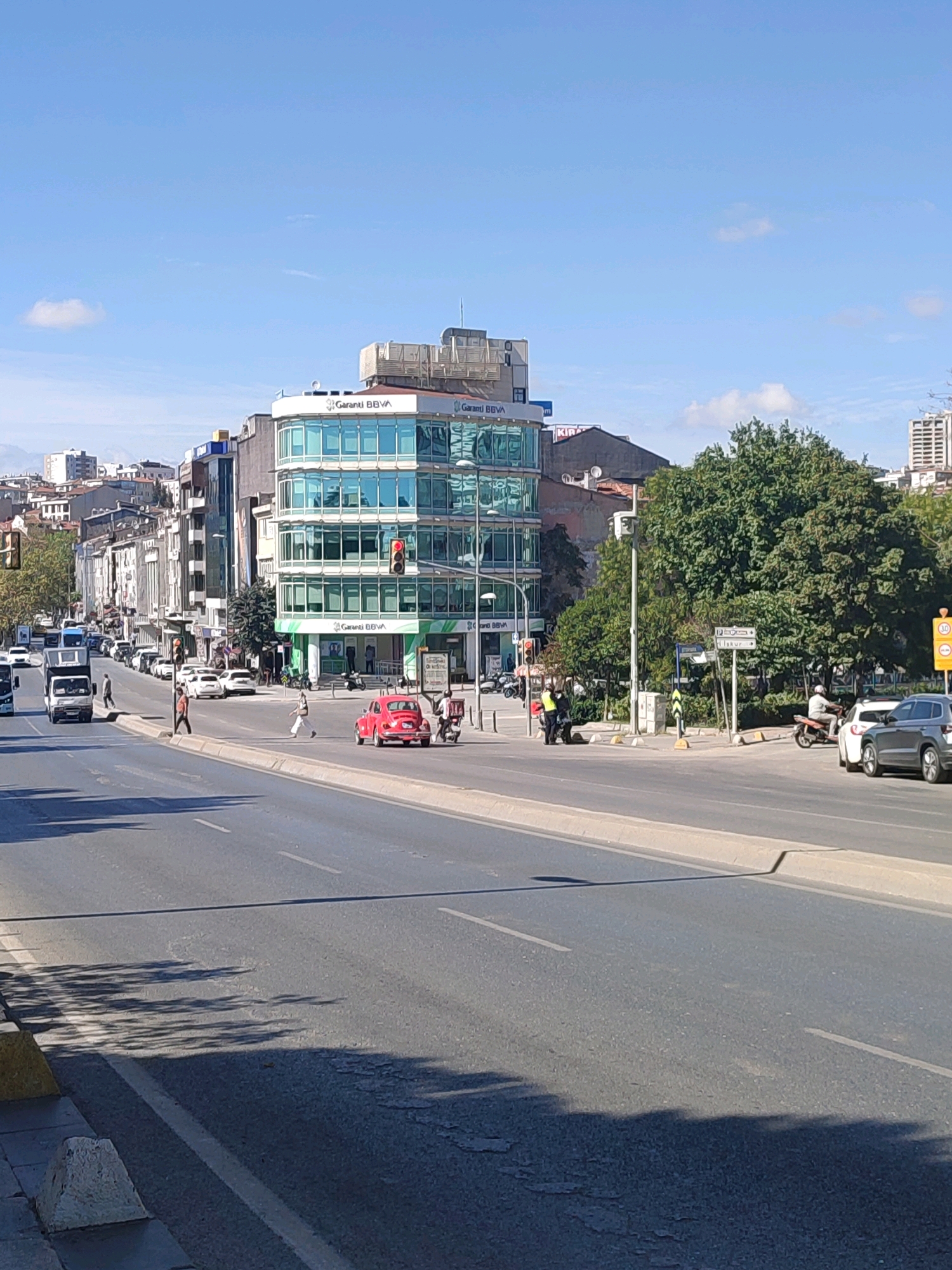 The image depicts a sunny day in Kadıköy, Turkey, focused on a wide, multi-lane road with moderate traffic. A modern, glass-fronted building, identified by "Garanti BBVA" signage, dominates the middle ground on the right side of the street. Across the road, on the left, is a line of older buildings. Several cars and a truck are visible on the road, with a red Volkswagen Beetle driving in the foreground. Two individuals in yellow vests appear to be directing traffic or working near the curb in the middle ground. Pedestrians are present on the sidewalks and crossing the street. Lush green trees are scattered throughout the scene, particularly behind the Garanti BBVA building, and shadows from trees are cast across the road. The sky is clear blue with a few wispy clouds. The overall impression is a snapshot of urban life in Kadıköy on a clear day, with a mix of contemporary and older architecture.