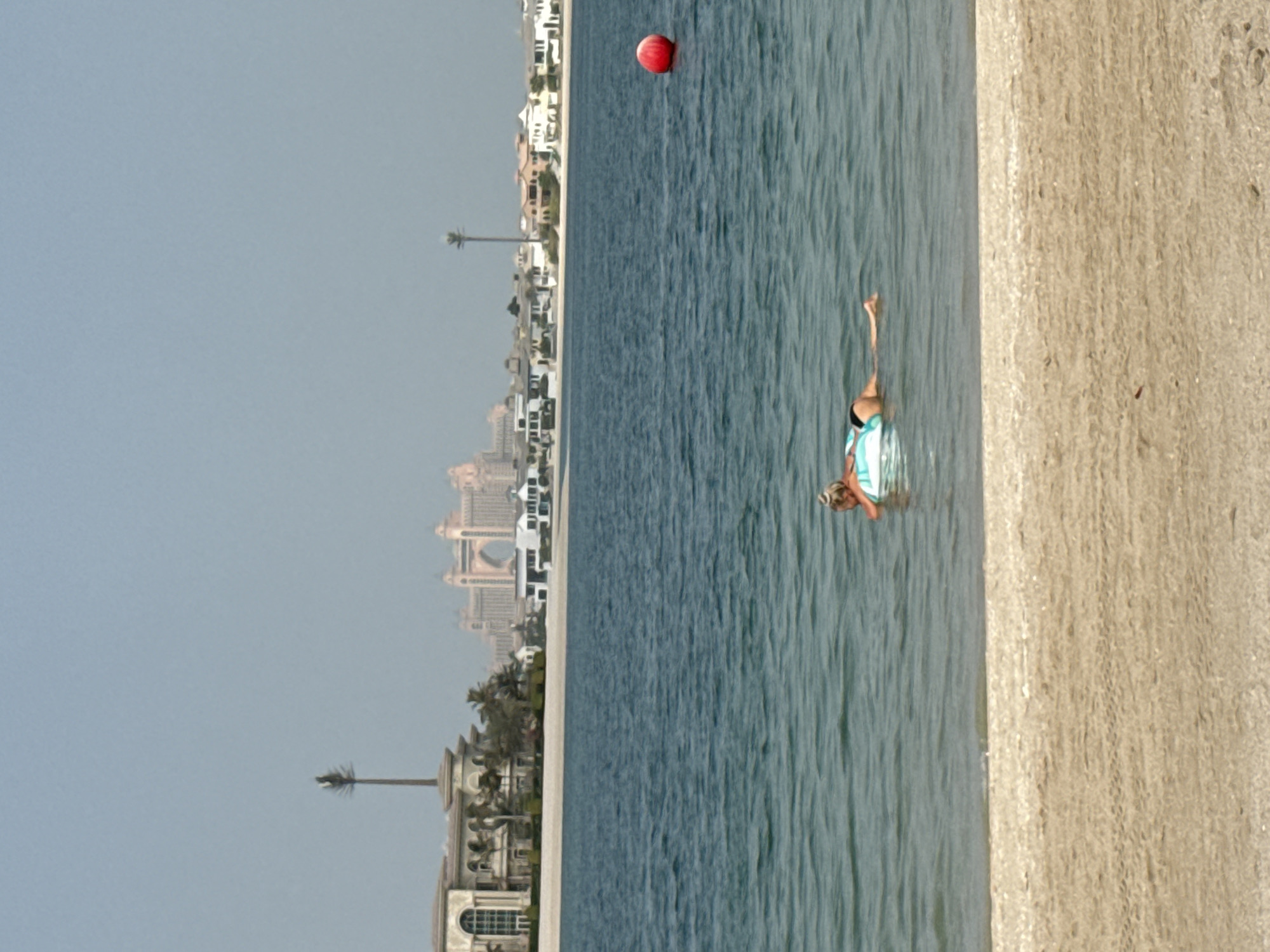 A medium shot shows a woman floating on an inflatable ring in the blue, choppy waters of the Persian Gulf, near a sandy beach. The woman, with blonde hair pulled into a bun, is facing away from the camera, her upper body is leaning back slightly. Her swimsuit is blue and white. In the distance, a cluster of upscale buildings lines the shore, with palm trees scattered amongst them. Further back, a large, arch-shaped hotel looms over the skyline. A single red buoy floats to the right of the frame. The sky is a hazy, pale blue. The image is likely taken from the Jumeirah area of Dubai, with the Atlantis, The Palm hotel visible in the background.