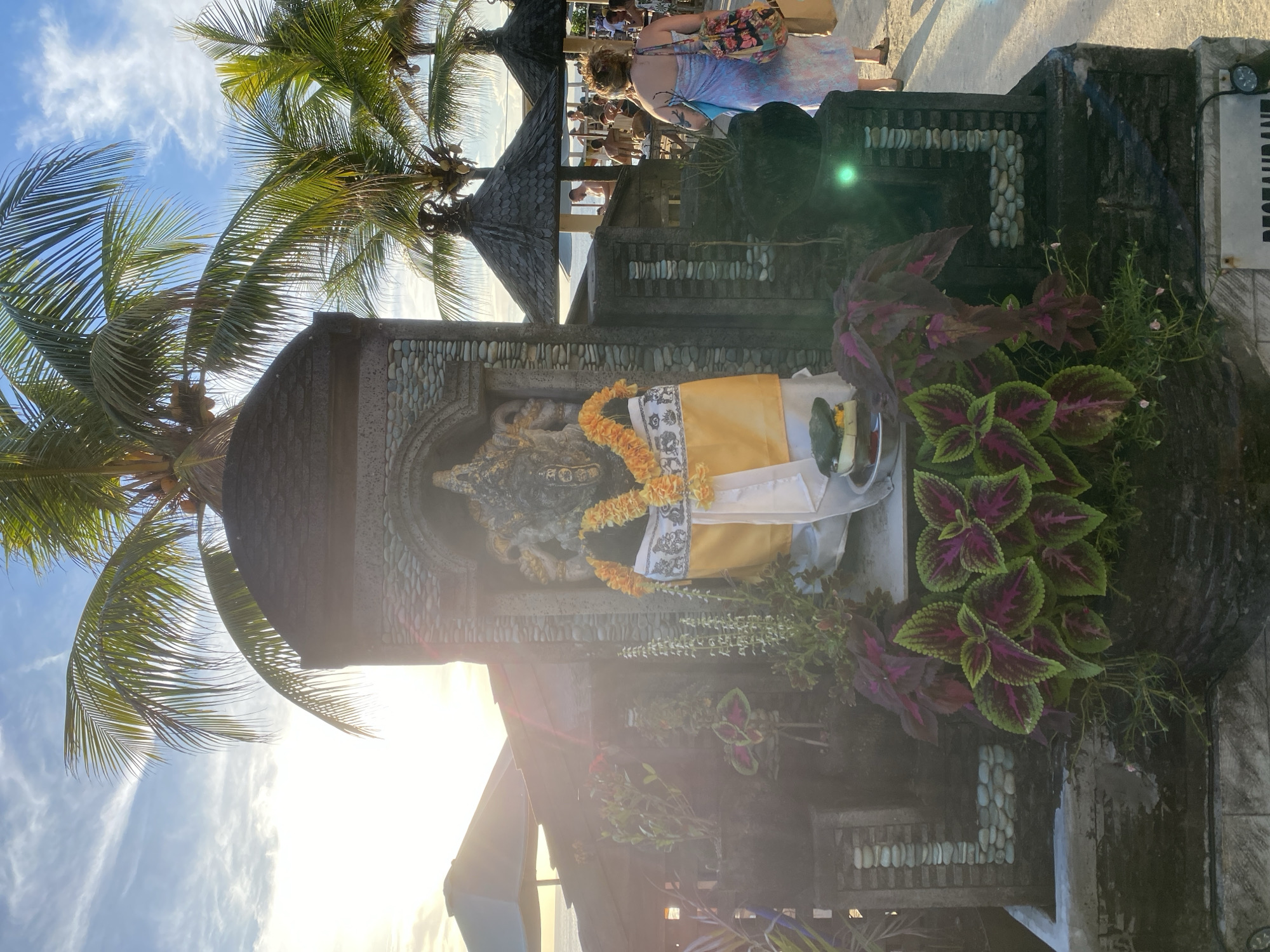 An outdoor scene in Kuta Selatan, Indonesia, captures a cultural shrine set against a tropical backdrop during late afternoon or early morning. The central subject is a stone statue, likely Ganesha, adorned with an orange-yellow garland and draped with yellow and white fabric. This statue is housed within a small, traditional structure with a dark, tiered roof, decorated with mosaic-like patterns of smooth, light-colored stones on its walls.
Around the shrine, various vibrant plants are visible, including coleus with striking green and purple leaves, and smaller flowering plants with tiny pink blossoms. Lush green palm trees with coconuts frame the scene, their fronds reaching towards a bright, partly cloudy sky. The sun is low, casting a strong, warm light that creates a prominent green and white lens flare in the image.
In the background, several people are gathered. Some are seen from behind, including a person wearing a floral shoulder bag and a light-colored dress/sarong. Others are seated at tables under small, dark-roofed pavilions, suggesting a restaurant or resort setting where people are dining or socializing.
The overall atmosphere is serene around the shrine, contrasting with the casual social activity in the background. The setting is distinctly tropical, characterized by the palm trees and traditional architecture. On the far right edge of the image, partially visible white text on a dark background reads "RESTAURAN," confirming the presence of an eatery nearby.
