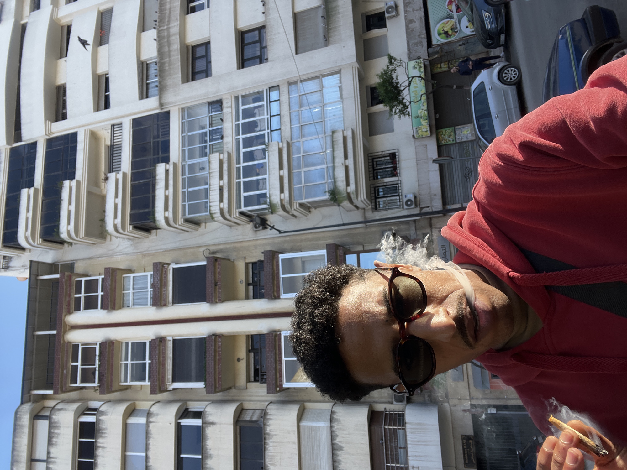 The image captures a male individual in the foreground, smoking a hand-rolled cigarette, set against an urban environment in Tanger, Morocco. The subject is a light-skinned male with dark, curly hair and a mustache, wearing dark, tortoiseshell-frame sunglasses and a red hooded sweatshirt. He holds the hand-rolled cigarette in his right hand, from which white smoke billows, and is actively exhaling smoke from his mouth. He is positioned close to the camera, looking forward and slightly downward.
The background consists of two multi-story urban buildings under a clear blue sky, suggesting daylight. The building immediately behind the subject on the left is a light-colored, possibly beige or grey, structure with several balconies, some of which are enclosed with windows and fitted with external blinds. The building on the right is a white, multi-story structure characterized by numerous glass-enclosed balconies and large windows. A black bird is visible in flight near an upper balcony of the white building, while other birds are perched on lower ledges.
At street level, below the buildings, a paved road hosts several parked vehicles, including a silver hatchback and a dark blue sedan. A second male individual, dressed in dark clothing, stands on the sidewalk adjacent to the silver car. Commercial storefronts are visible at the ground level of the buildings, one displaying Arabic text and an illustration of produce. The scene depicts a typical street in an urban district.