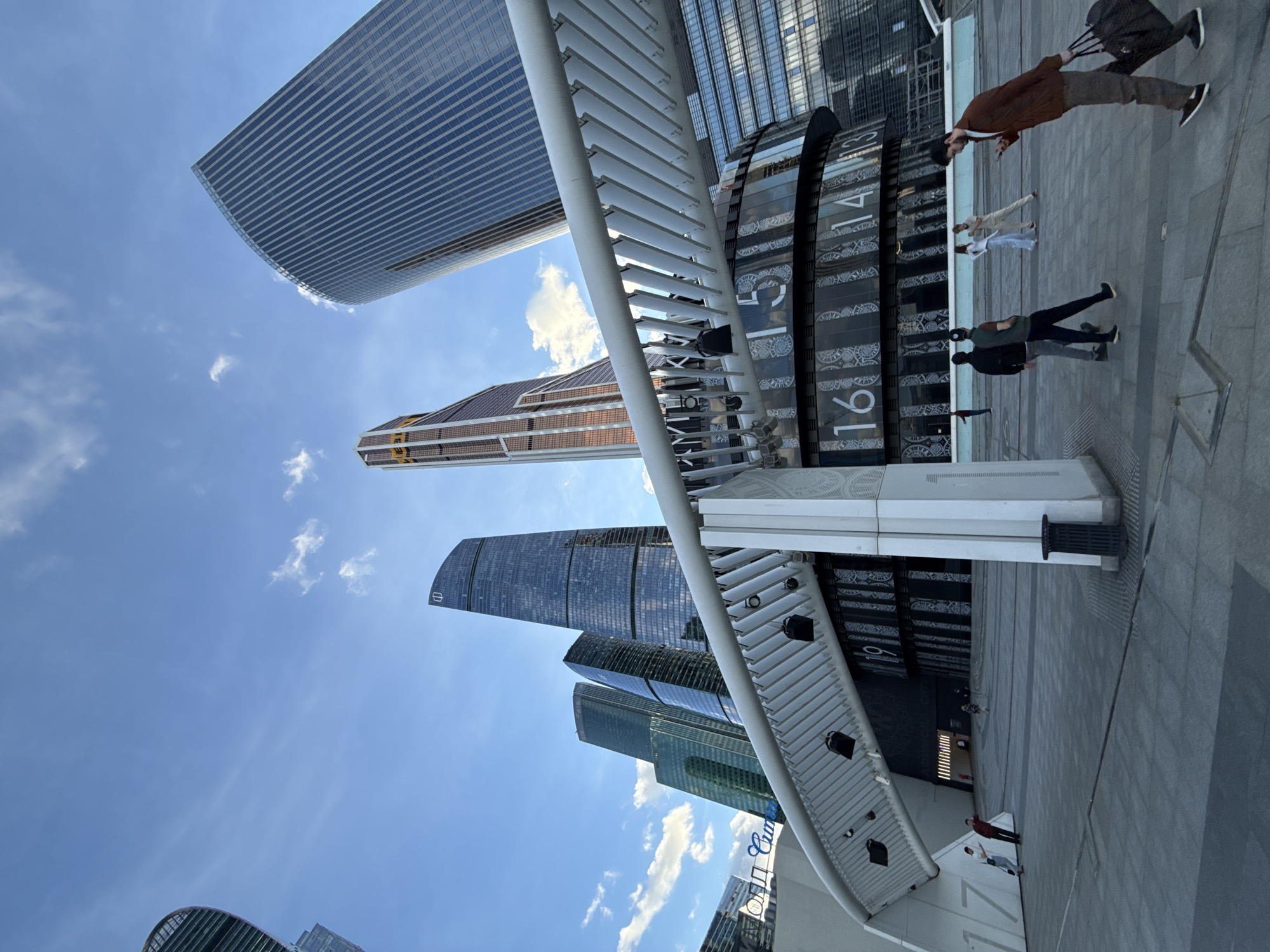 This media file showcases a scene in Moscow, Russia, featuring modern skyscrapers under a bright blue sky with scattered white clouds, suggesting a daytime setting. In the foreground, a unique cylindrical clock with large Roman numerals is visible. Several people are walking along a paved area near the clock, with some carrying bags, indicating they are travelers or commuters.
One individual, dressed in a brown jacket and grey pants, is seen walking away from the camera, pulling a dark suitcase. Another person in a light-colored outfit walks in the same direction, while a third individual in a dark jacket and cap is walking towards the camera. The architecture of the surrounding buildings is characterized by glass facades and sharp, modern lines.
The overall atmosphere appears calm and busy, with the people engaged in everyday activities. The presence of the large clock and the imposing skyscrapers suggests an urban, possibly commercial or business district. The clear sky and sunshine point to favorable weather conditions for outdoor activities. Some text is faintly visible on the clock face and on a sign in the background, but it is not legible enough to be read. The image is oriented vertically, with the sky at the top and the ground at the bottom.