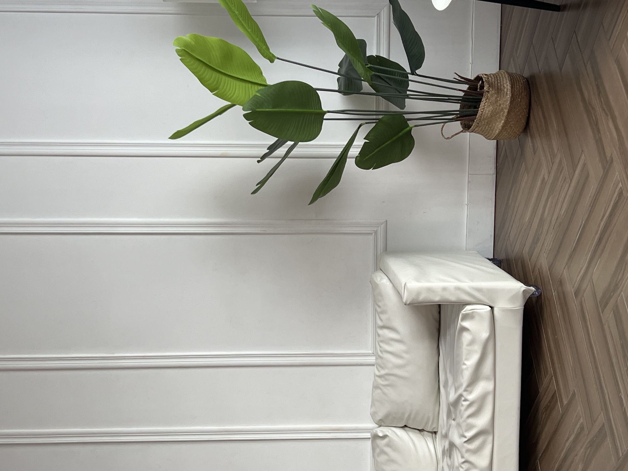 The image shows an interior space with a white wall decorated with molding on the left side. To the right is a white sofa with multiple cushions. Adjacent to the sofa is a portion of a brown, herringbone-patterned floor. In the upper portion of the image, a potted plant with large green leaves sits in a woven basket. The location is consistent with an indoor space, but there is no information to confirm the location as the city of Chaikovsky, Russia.