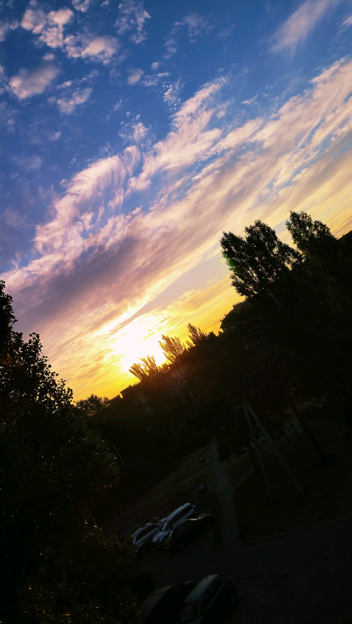 The image captures a sky filled with wispy clouds painted in shades of blue, pink, and yellow, indicative of a sunset or sunrise. The sun itself is a bright, luminous orb, casting a warm glow across the horizon. Silhouetted against this vibrant sky are the dark shapes of trees and buildings, suggesting an urban or suburban setting. In the foreground, several cars are parked on what appears to be a paved surface, possibly a street or parking lot. The overall scene is a snapshot of a tranquil evening in the city of Horishni Plavni, Ukraine.