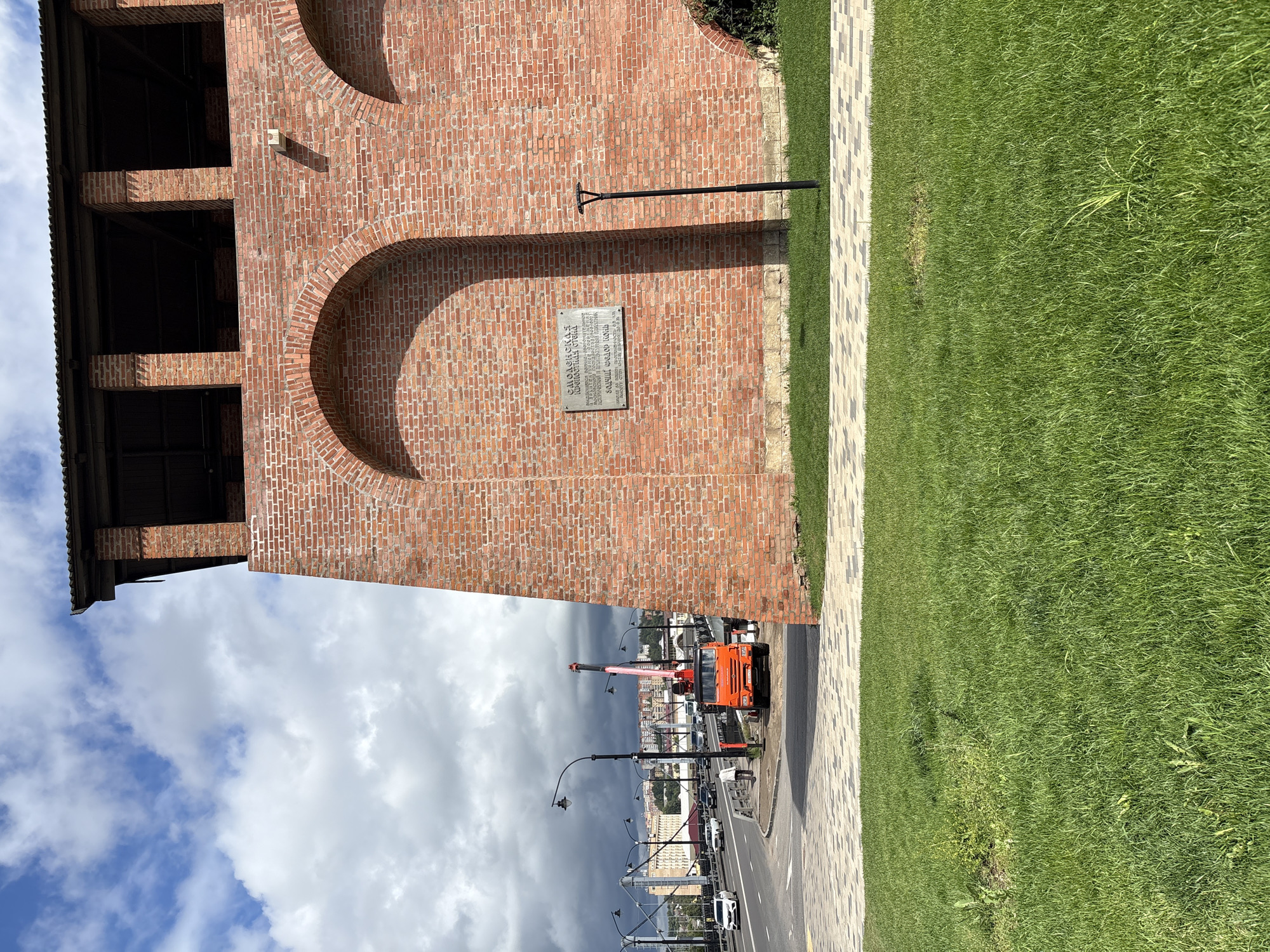 The image shows a close-up view of a section of a large brick wall with arched openings, likely part of a historical fortification or building. The wall is made of red bricks and has a dark wooden roof structure at the top. A plaque is affixed to the wall, bearing text that is difficult to read clearly. To the right of the plaque, a modern, black, minimalist lamppost stands on a patch of green grass.
In the background, a road with several cars is visible, including a white sedan and an orange truck with a crane attached. Streetlights line the road, and a large bridge with a metal structure can be seen in the distance. The sky is partly cloudy with some dark storm clouds gathering.
The scene appears to be an urban environment with a mix of historical architecture and modern infrastructure. The presence of the truck with a crane suggests ongoing construction or maintenance work. The specific location mentioned, Demenshchina, Russia, would imply this is a view of a landmark or historical site within that city.