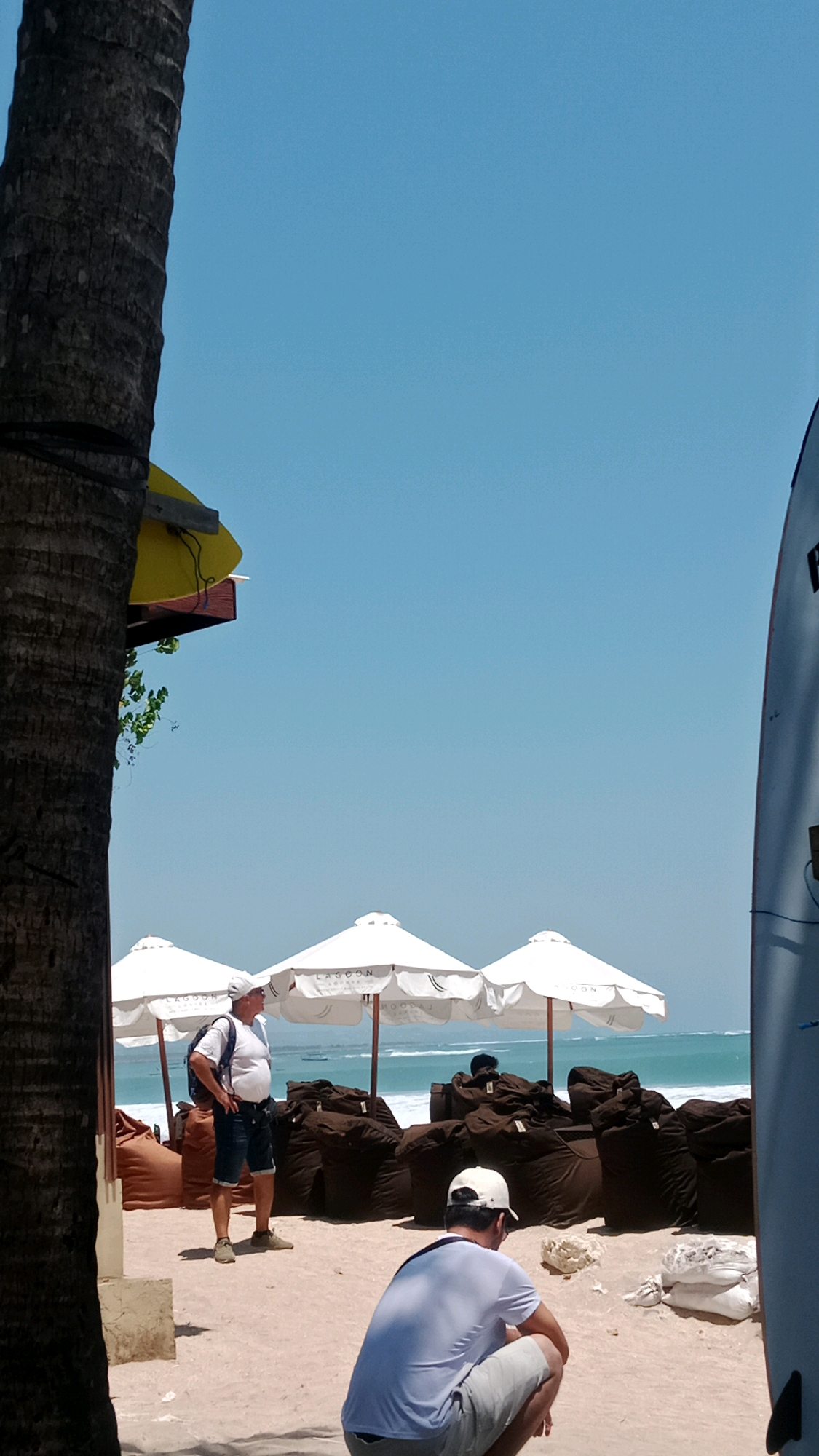 An outdoor shot captures a beach scene in Kuta, Indonesia, under a clear blue sky. A large palm tree trunk dominates the left foreground, partially obscuring the view of the sandy beach. In the middle ground, three white beach umbrellas with the word "LAGOON" printed on them are set up, shading numerous dark brown beanbag chairs.
Two men are present in the scene. One man, wearing a white t-shirt, blue denim shorts, a white baseball cap, and a backpack, stands in the middle of the beach with his hands on his hips, looking out towards the ocean. Another man, wearing a white t-shirt and light-colored shorts, crouches on the sand in the right foreground, facing away from the camera.
The ocean is visible in the background, with gentle waves and a few distant boats. The sand is light in color and appears to be a typical beach setting. A tall, light-colored surfboard stands upright on the far right of the frame. A yellow object is visible attached to the palm tree trunk, possibly a sign or decoration.