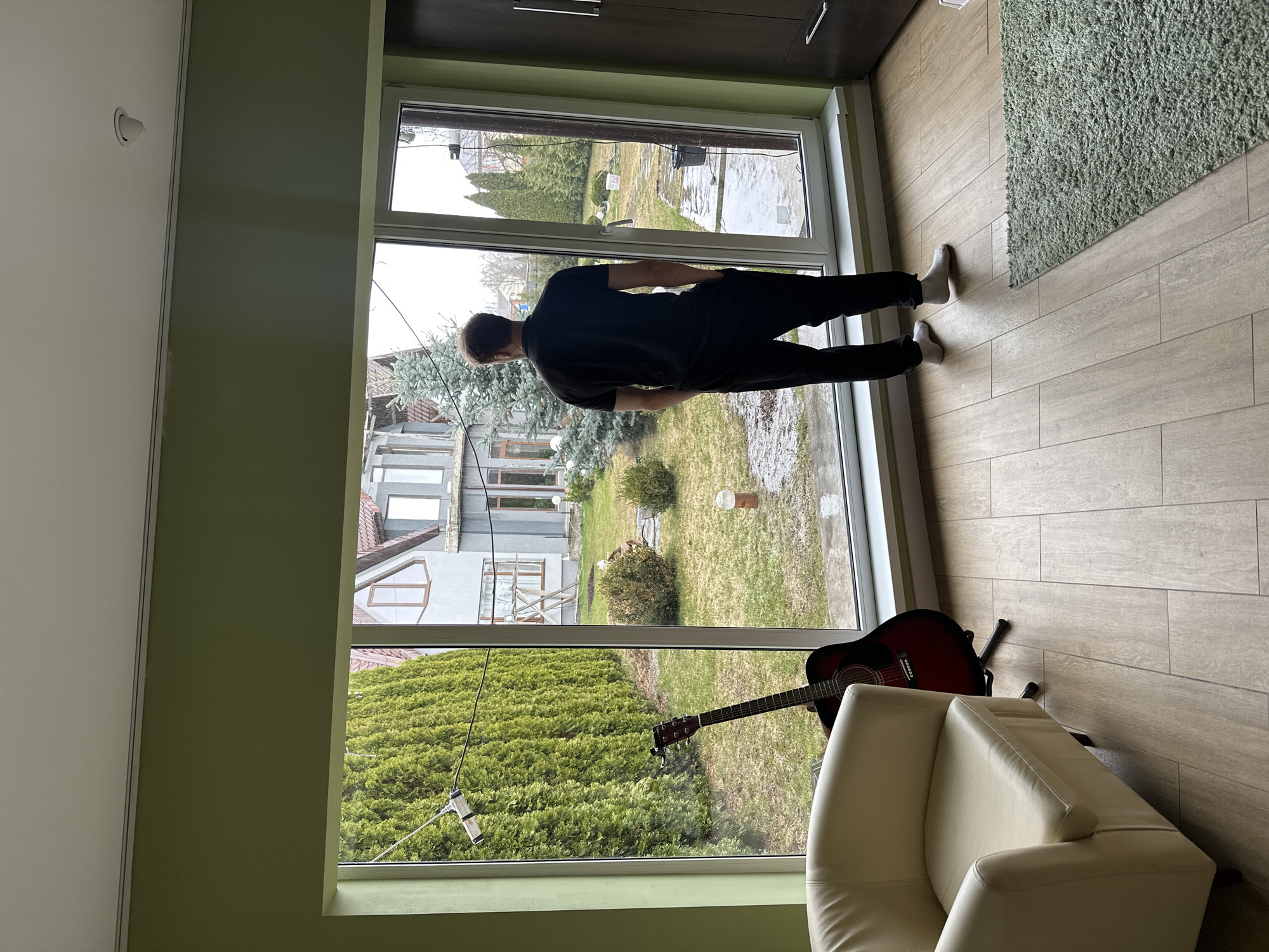 A man in a black t-shirt and black pants stands in front of a large window, his back to the camera. He is wearing white socks. He appears to be looking out at a green lawn and houses. Outside, the ground is wet, possibly from rain. Indoors, a white chair sits in the lower right corner, and a red guitar rests on the floor by the chair and the window. There is a green rug in the upper right corner and wood-look tile flooring. The room has green walls. A window cleaning tool rests against the window. The photo does not provide definitive location in Kursk, Russia.