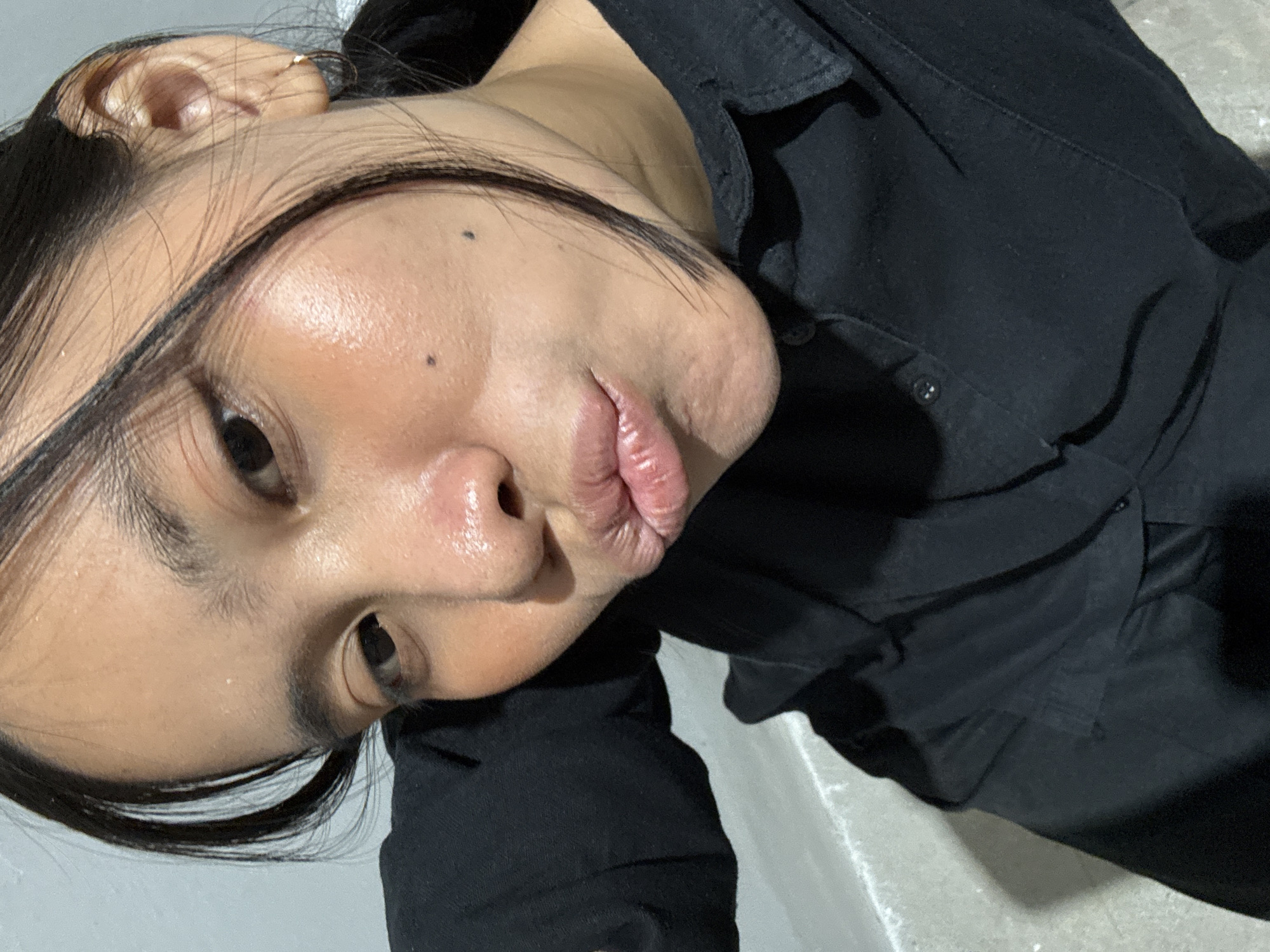 The image shows a close-up selfie of a young woman with dark hair and light skin. She is wearing a black collared shirt. Her face is angled towards the camera, and her lips are puckered. There are a few small dark moles visible on her cheeks. The background appears to be a light-colored wall and a greyish floor. The lighting suggests an indoor environment. The metadata for this image does not contain any information about the location in South Jakarta, Indonesia.