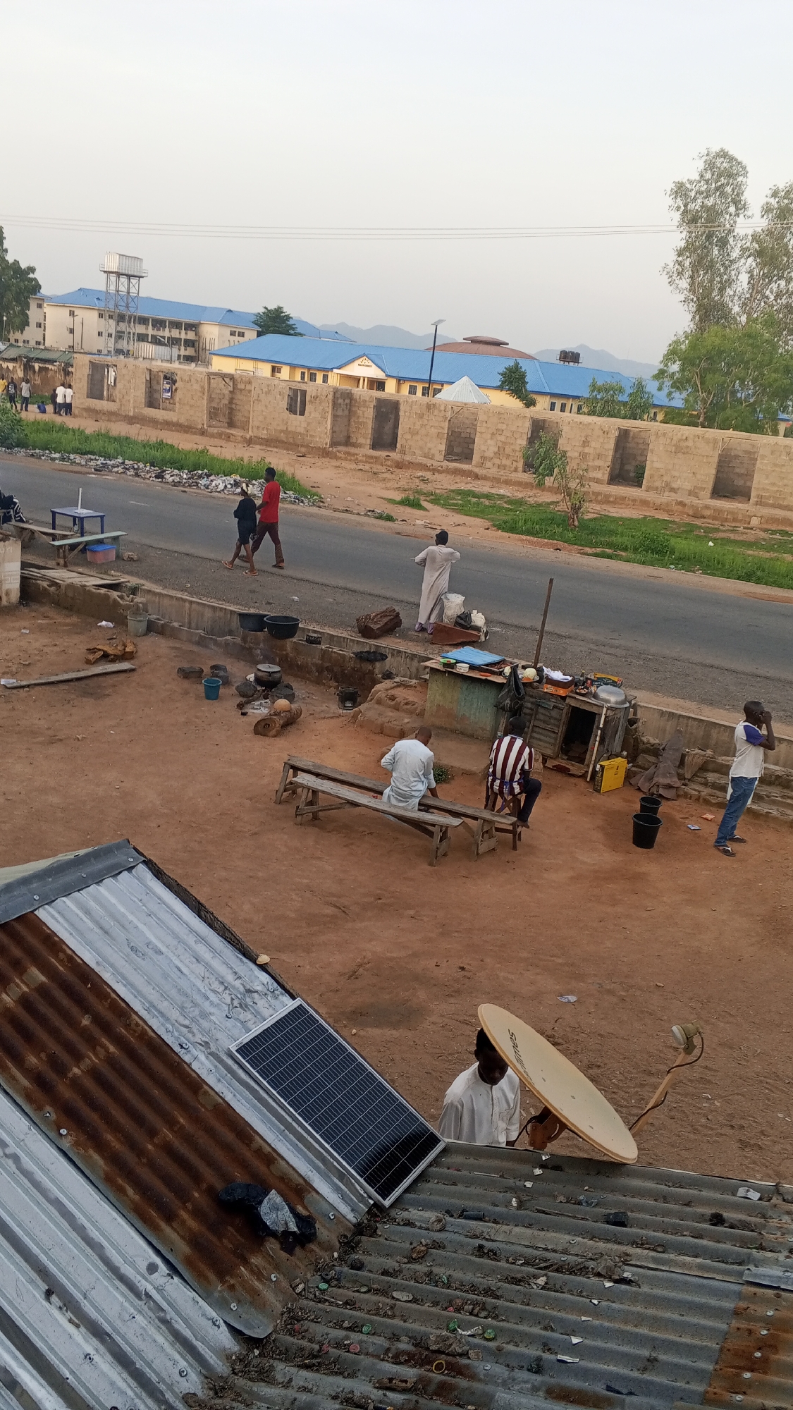 The image captures a street scene in an urban environment. In the background, a large building complex with blue roofs is visible, featuring a water tower. In the middle ground, a road runs parallel to a construction site where brick walls are being built. Several individuals are walking on the road and along the sidewalk. The foreground shows a dusty, open area with several people sitting on benches and a small stall with various items. There are cooking pots and firewood in this area, suggesting food preparation. A solar panel is installed on a corrugated metal roof in the lower left of the frame, and a satellite dish is mounted on a similar roof to the right, with a person standing nearby. The overall scene depicts a blend of residential, commercial, and construction activities in what appears to be a developing area.