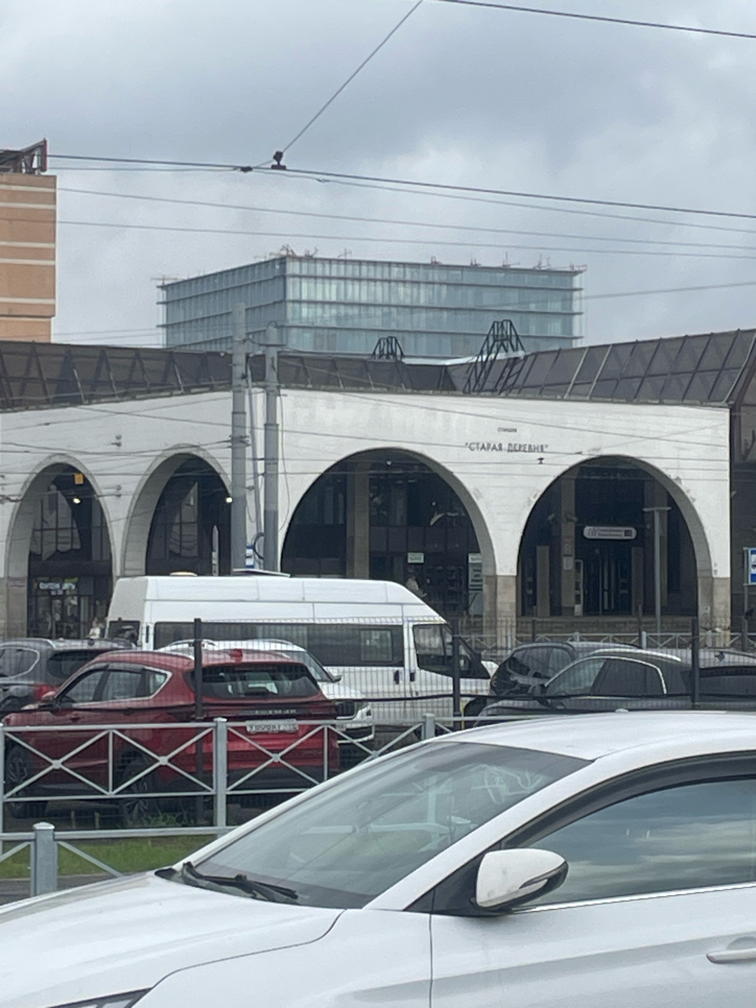 The image shows a cloudy day in Saint Petersburg, Russia. In the foreground, the hood and side mirror of a white car are visible. Behind it, a red SUV is parked behind a metal fence. Further back, a white van is parked in front of a building with arched entrances. Above the building, a modern glass structure is visible in the distance. The building has a sign that reads "СТАРАЯ ДЕРЕВНЯ" (Staraya Derevnya).