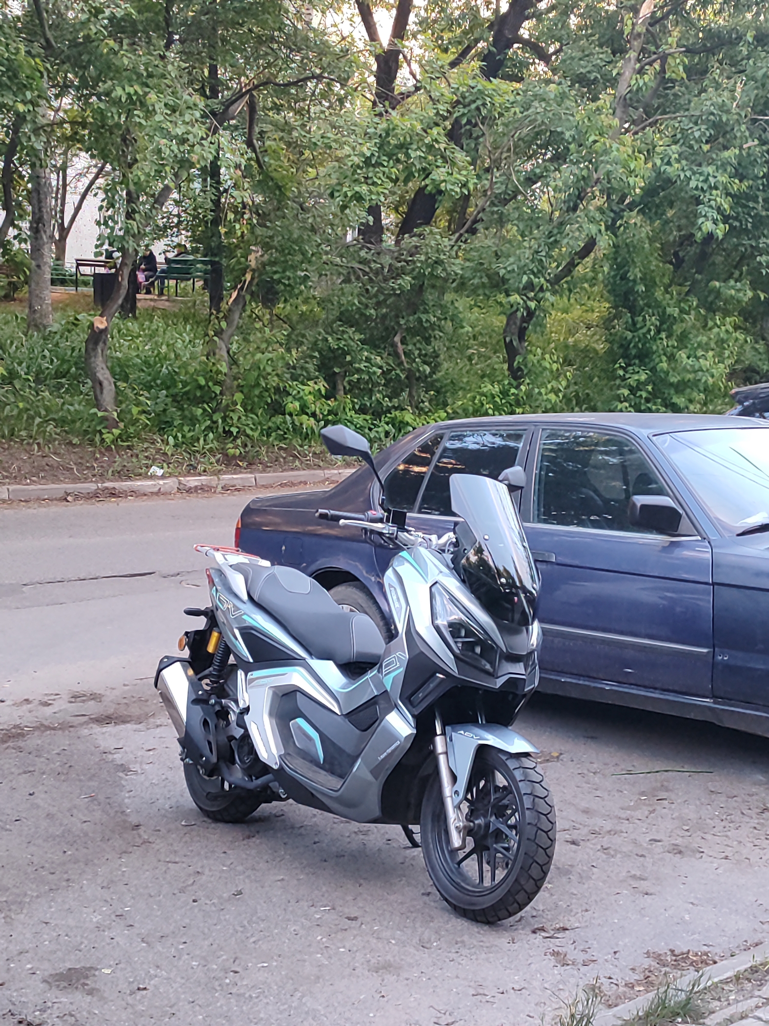 The image shows a gray Honda ADV scooter parked on a paved surface. A dark blue sedan is parked behind the scooter. In the background are trees and a bench with people seated on it. The location is likely Shchyolkovo, Russia, though this is based on image metadata not visual details.