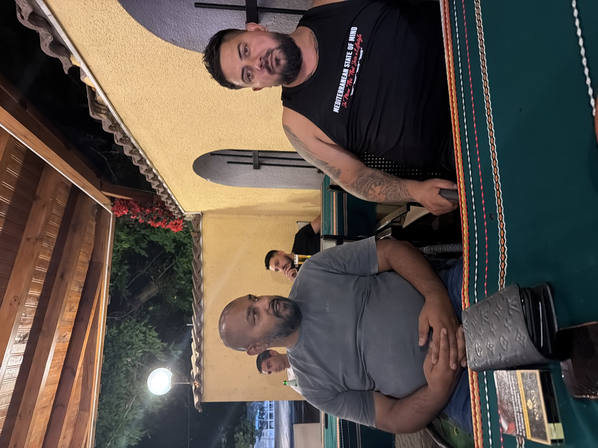 The image shows two men sitting at a table at an outdoor restaurant. The man on the left is bald with a beard and is wearing a gray t-shirt. The man on the right is wearing a black tank top with text that reads "MEDITERRANEAN STATE OF MIND". Both men appear to be middle-aged. There are other people visible in the background. On the table in front of the man on the left, there is a wallet and a pack of cigarettes. The restaurant appears to be located in the city of Levski, Bulgaria, based on the surrounding architecture which includes a yellow stucco building with arched doorways and a wooden ceiling. The lighting suggests it is evening.