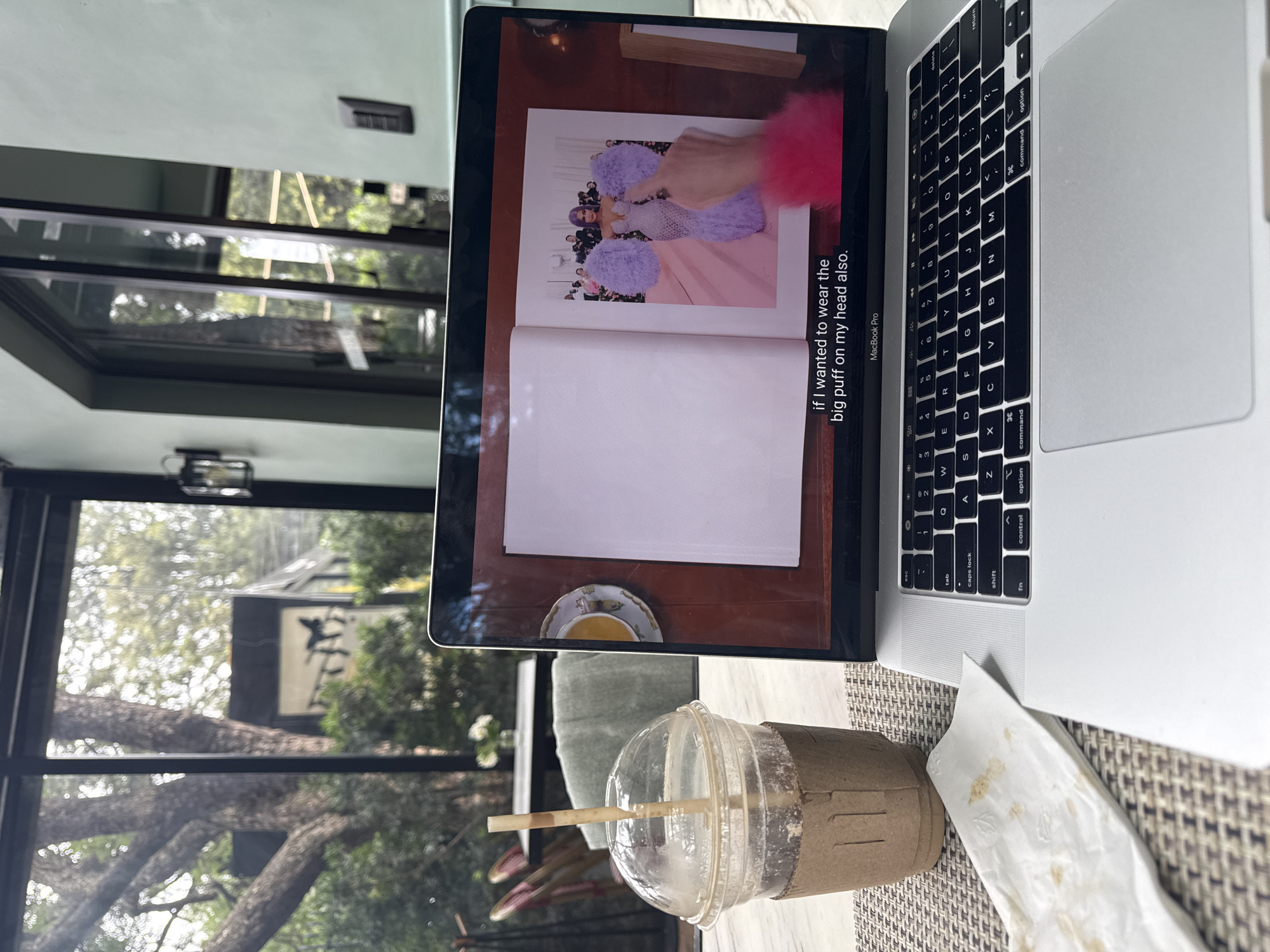 A close-up, eye-level shot captures a laptop screen displaying an open book with a fashion photo and text overlay, positioned on a textured placemat next to a partially consumed iced coffee in a plastic cup with a straw and a brown cardboard sleeve. A crumpled white napkin with brown stains rests on the placemat to the left of the coffee cup. The laptop keyboard is visible in the foreground. Through the laptop screen and a glass wall in the background, an outdoor scene with trees, foliage, and outdoor seating furniture is discernible, suggesting the location is a cafe or restaurant with an indoor-outdoor seating area. The lighting appears to be natural, with some artificial light sources visible in the background as sconces on a pale green wall. The text on the laptop screen reads, "if I wanted to wear the big puff on my head also." The overall setting suggests a relaxed, contemporary atmosphere, possibly in a cafe or restaurant in Mexico City, Mexico, where someone is engaged with content on their laptop.