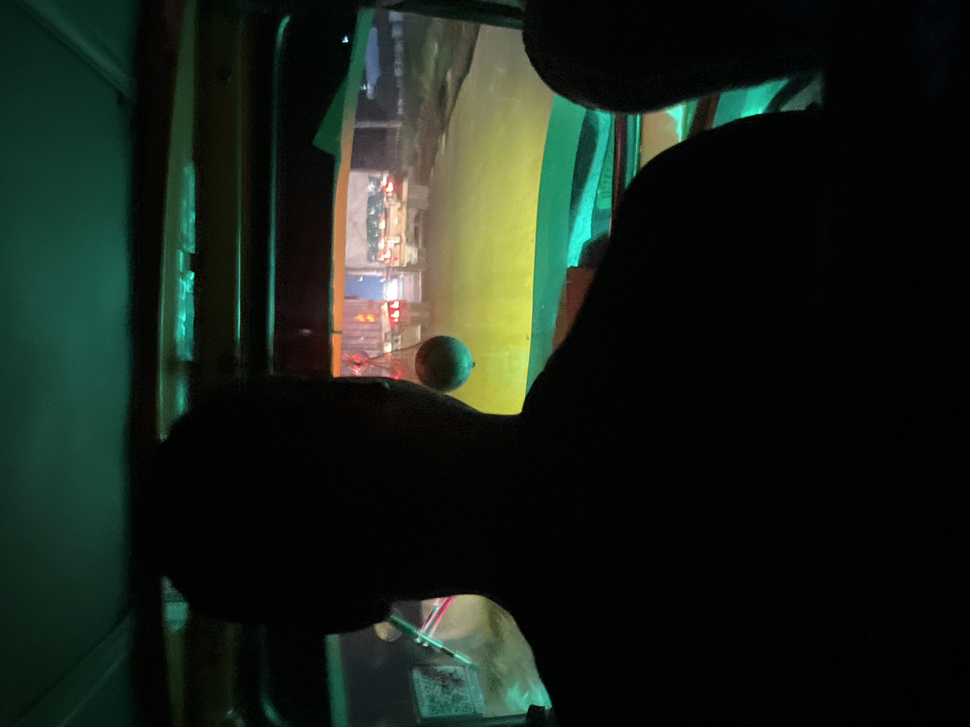The image depicts the interior perspective from a vehicle, likely at night, in the city of Unknown, Unknown. A person, seen from behind in silhouette, occupies the central foreground, presumably the driver or front passenger. Another partial silhouette is visible on the far right. The vehicle's interior roof and frame are illuminated by a green light source. A yellowish-green light emanates from the lower front of the vehicle, illuminating the immediate road ahead. Suspended from the upper interior, a small, spherical object hangs to the left of the central person's head. Outside the vehicle, two large trucks are visible directly ahead, displaying illuminated red taillights. The road surface appears paved. The background is dark, with indistinct shapes suggesting distant structures or vegetation under the night sky.