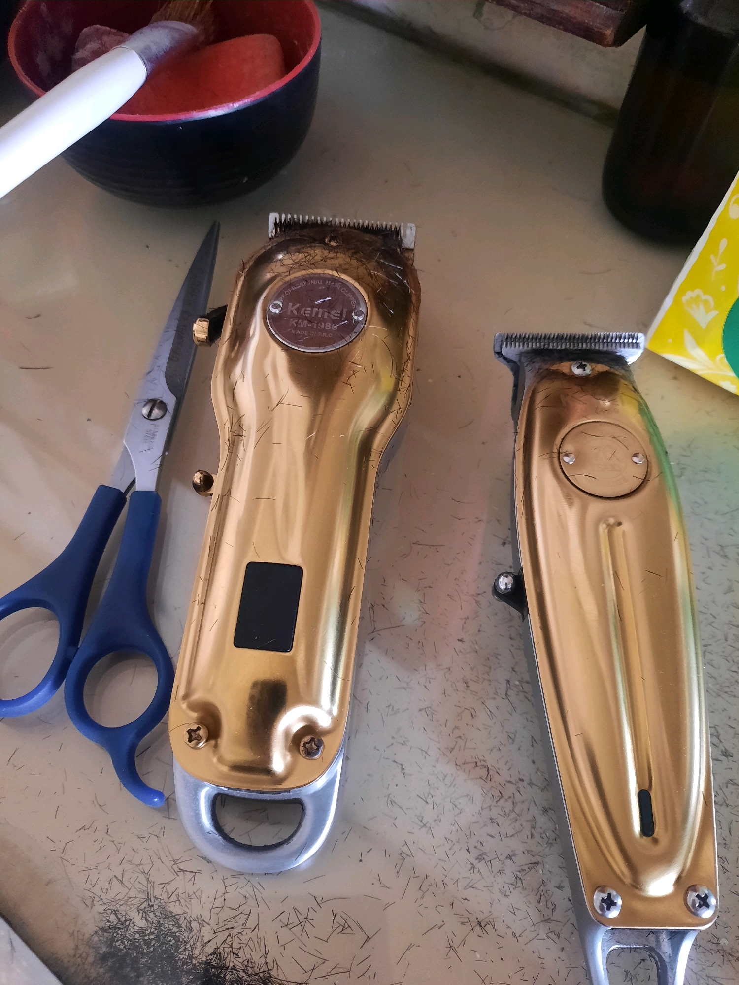 The image depicts a close-up, overhead view of a collection of barbershop tools on a light-colored, textured surface strewn with hair clippings. Two golden hair clippers, one larger than the other, are prominently featured. The larger clipper, with a visible "Kemei KM-1986" marking, shows signs of use, with hair caught in its blades. A pair of blue-handled scissors lies to the left of the larger clipper. In the background, a black bowl with a red rim contains a white-handled brush and a red sponge-like object. A dark brown bottle and a portion of yellow packaging with floral motifs are visible in the upper right corner. The scene suggests a recent haircutting session. The location is identified as East Jakarta, Indonesia.