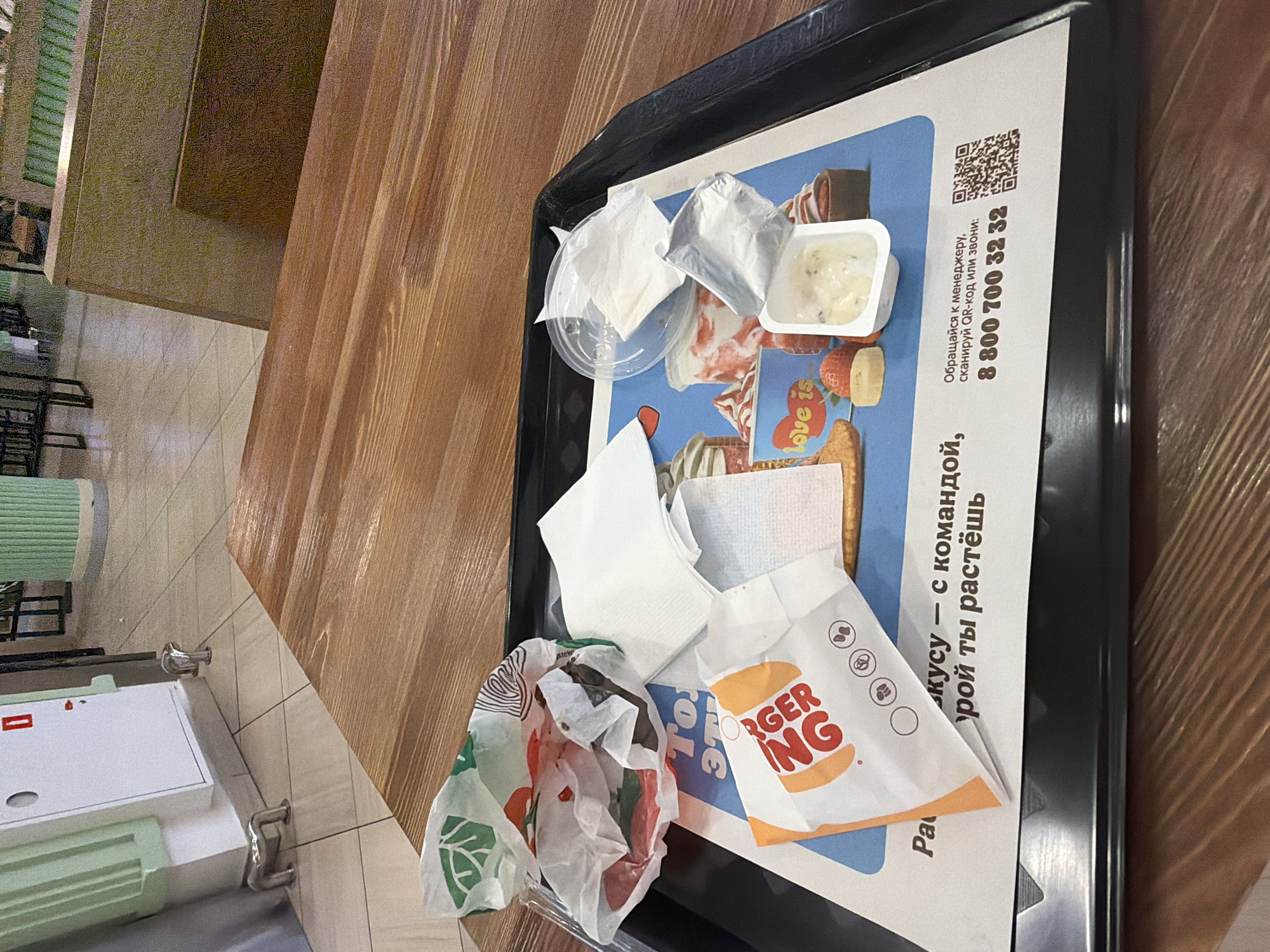 This media file shows a close-up, overhead view of a Burger King tray on a wooden table. The tray contains used fast-food wrappers and a small container of sauce. A white paper wrapper with the Burger King logo and branding is prominently displayed. There are also crumpled paper napkins and a clear plastic lid with some napkins inside. A QR code and a phone number, "8 800 700 32 32", are visible on the tray liner, along with some Russian text that appears to be related to customer service. The setting appears to be indoors, likely a Burger King restaurant, given the branding. The lighting suggests it could be daytime. There are no people visible in the image, and the focus is on the discarded food items and the tray itself. The overall impression is one of a recently finished meal.