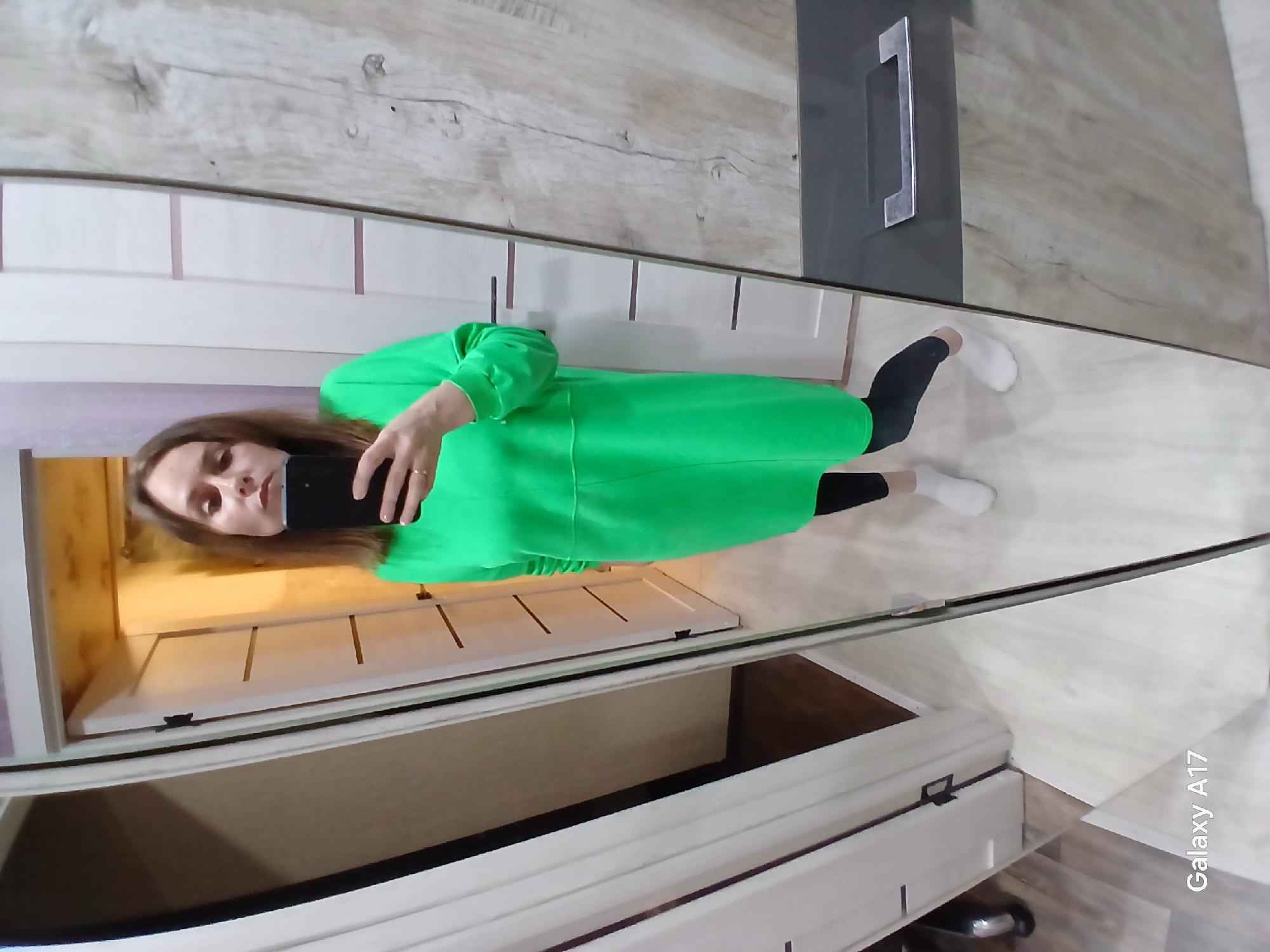 A woman with shoulder-length brown hair is taking a selfie in a full-length mirror. She is wearing a bright green, long-sleeved, midi-length dress, black leggings, and white socks. She is holding a black smartphone in front of her face.
The mirror is reflecting a room with light-colored wood grain laminate flooring and furniture. Some of the furniture appears to be white with horizontal recessed lines. A partial reflection of a cupboard door with a grey panel and a silver handle is visible. The bottom portion of the mirror's reflection shows drawer fronts.
The photo includes a watermark indicating "Galaxy A17." The location of the image is implied to be in Yekaterinburg, Russia, but this is not directly depicted in the image.