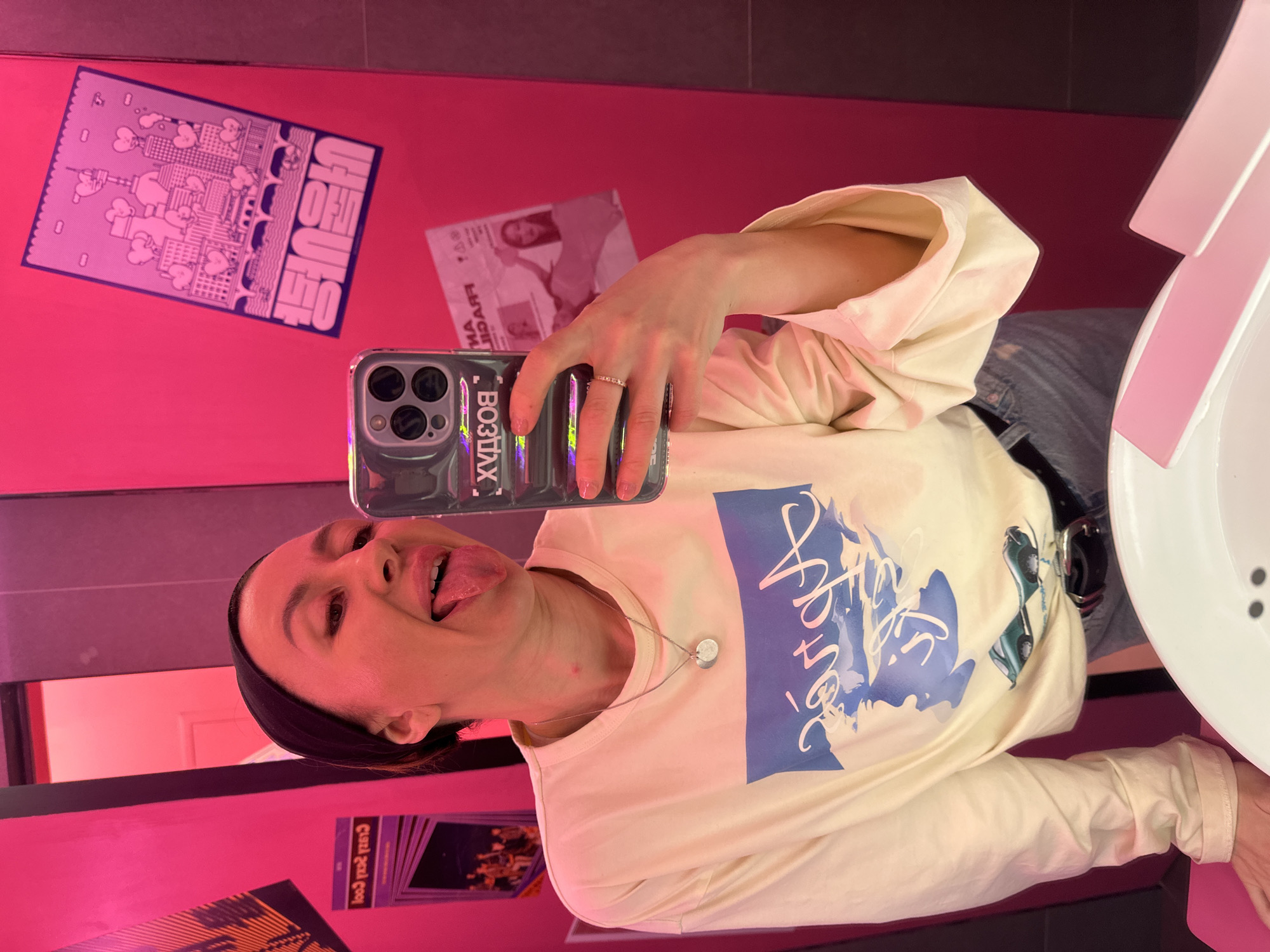 A young woman takes a mirror selfie in a vibrant, pink-walled room, likely a restroom or changing area. She stands vertically, facing the camera with her tongue playfully sticking out. She wears a cream-colored long-sleeved t-shirt with a large blue graphic print on the front, blue jeans, and a black headband. A silver necklace with two small circular pendants hangs around her neck, and a ring is visible on her right ring finger. She holds an iPhone in a holographic/iridescent clear case featuring three camera lenses.
The background is dominated by bright pink walls. To the right, a white sink with pink accents is partially visible. The walls are adorned with several posters. On the upper left, a light purple poster depicts a cityscape with hearts and stylized buildings, featuring the text "UEGUH" (possibly stylized and mirrored). Below that, another paper or poster shows a face (likely a woman's) with partially visible mirrored text, including "МА ДИЯАЧI". On the bottom left, a dark poster with orange/yellow graphics contains mirrored text "СУЛ 2оол". The text visible on her phone case, reflected in the mirror, reads "ХУЗДОВ", which is the mirrored version of "ВОЗДУХ" (VOZDUKH, meaning "air" in Russian). The blue graphic on her shirt is a stylized script that is difficult to decipher clearly. The setting is brightly lit, creating a cheerful atmosphere.