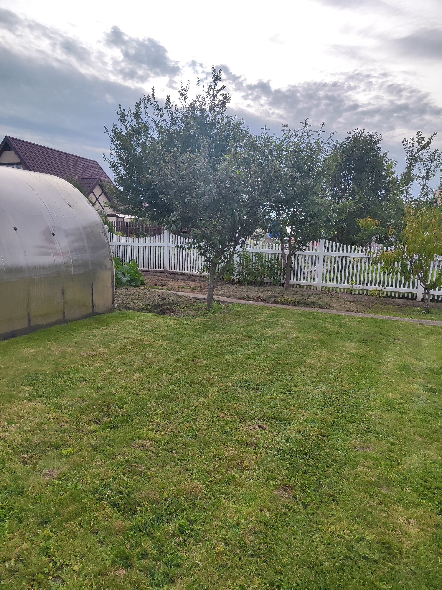 The image depicts a backyard scene in Gvardeysk, Russia. In the foreground, a well-maintained green lawn fills the lower half of the frame. To the left, a large, translucent greenhouse is partially visible. Behind the greenhouse, a portion of a house with a dark red, sloped roof and decorative trim is present. A white picket fence runs horizontally across the middle ground, separating the lawn from a more cultivated area. Beyond the fence, various trees with green foliage are visible, along with what appears to be a garden or raised beds. The sky above is overcast with scattered clouds. The scene suggests a residential property with gardening and outdoor structures.