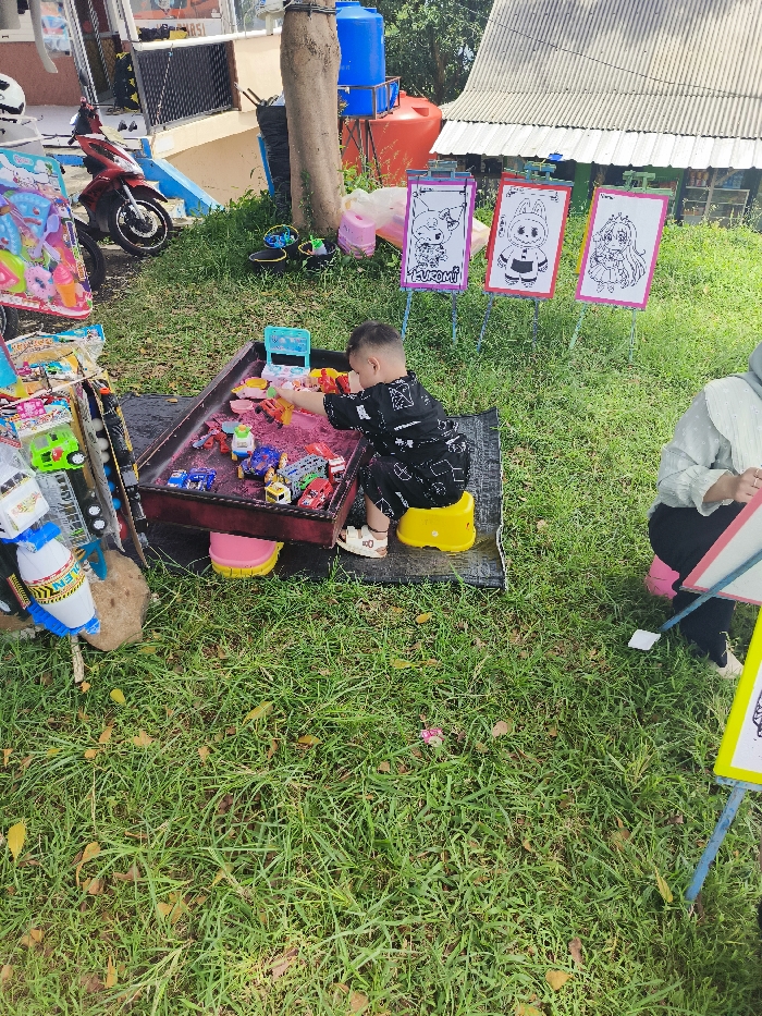 The scene is set in a grassy area with a sandpit play area. A young boy, wearing a black and white shirt and shorts, sits on a yellow bucket and plays with a toy car in the sandpit. The sandpit is made of wood and has pink plastic lining, with a small toy cash register and several other plastic toys. The boy is wearing a black backpack. Behind the sandpit, on the ground, are three wooden easels holding drawings of cartoon characters on paper. There is a person in the background wearing a white headscarf and holding a white notepad. Behind the sandpit are several other items such as a toy motorcycle and a blue water tank. The scene is located in Majalengka, Indonesia. The coordinates provided are -6.846 latitude and 108.204 longitude.
