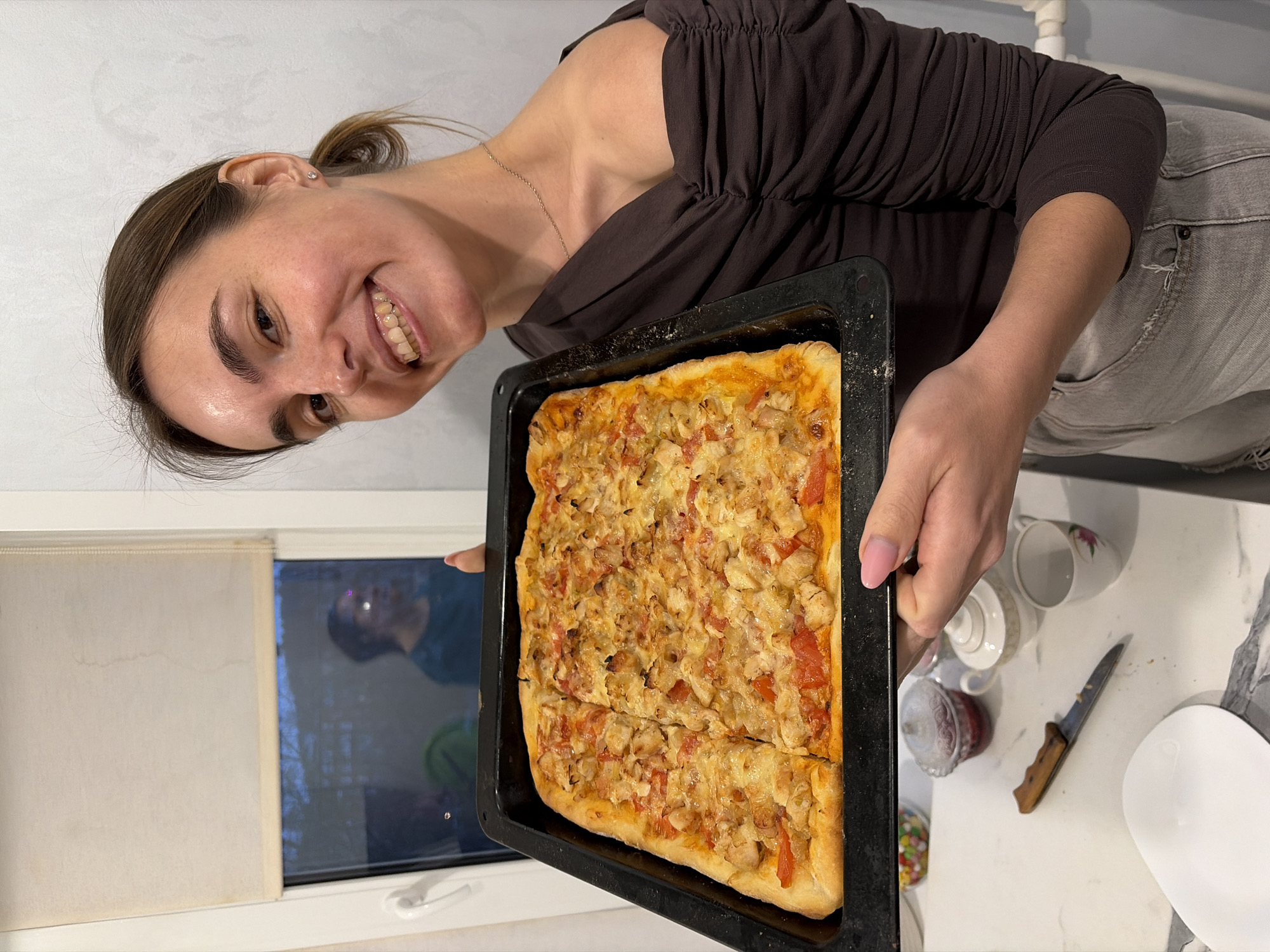 A woman is holding a rectangular baking sheet containing a rectangular pizza, presumably homemade, in a domestic setting, likely a kitchen. The pizza appears to be cut into segments. The woman is smiling and looking at the camera.
Behind her is a window with partially drawn shades. A reflection is visible in the window. To the side is a countertop with cups, a knife, a white plate, and what appears to be candy. The woman is wearing a dark brown top and grey jeans. The picture appears to have been taken indoors. There are no visible indicators relating to Sarapul, Russia.