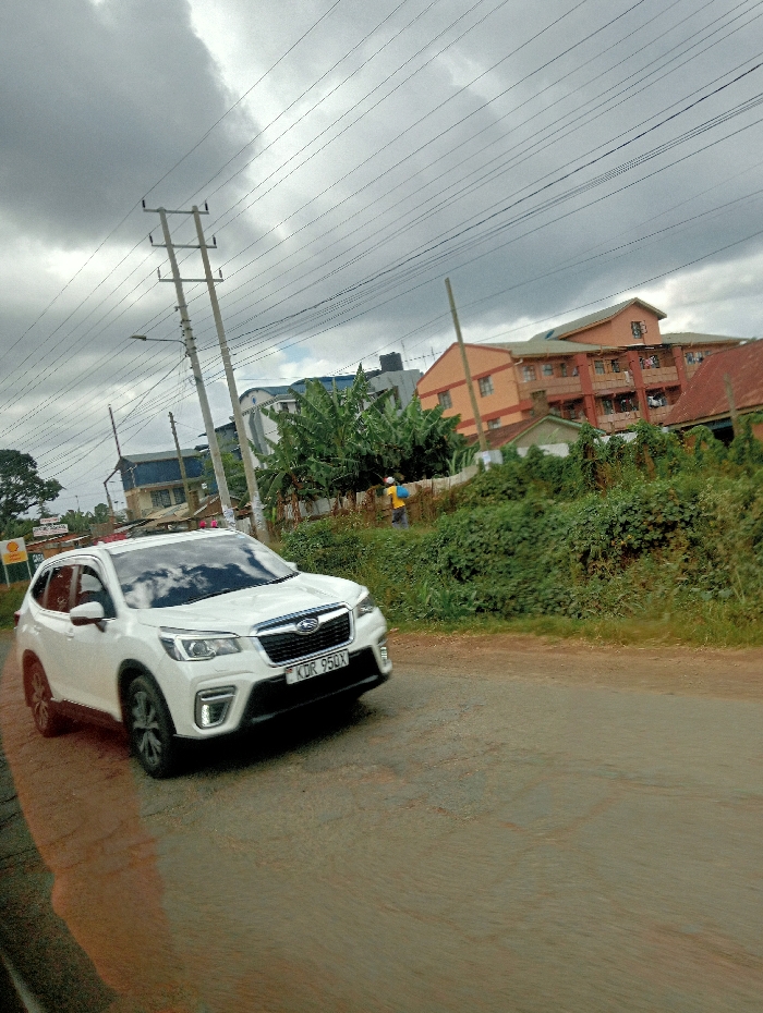 The image depicts a street scene in Karatina, Kenya, under an overcast sky. A white Subaru Forester SUV, bearing Kenyan license plate KDR 950X, is the prominent subject, positioned on a partially unpaved or degraded asphalt road, angled towards the right and appearing to be in motion. To the far left, a partial view of a green and yellow Shell gas station sign is visible. The mid-ground features a tall utility pole with numerous overhead power lines and an attached street light, flanked by dense green vegetation, including banana plants. Behind this foliage and pole, several multi-story buildings are visible; one features light blue roofing, while another prominent building has orange-colored walls and a reddish-brown tiled roof with clothes drying on balconies. A single individual wearing a yellow top and dark trousers is partially visible standing within the vegetation in the middle-ground. The sky above is uniformly grey with dense clouds.