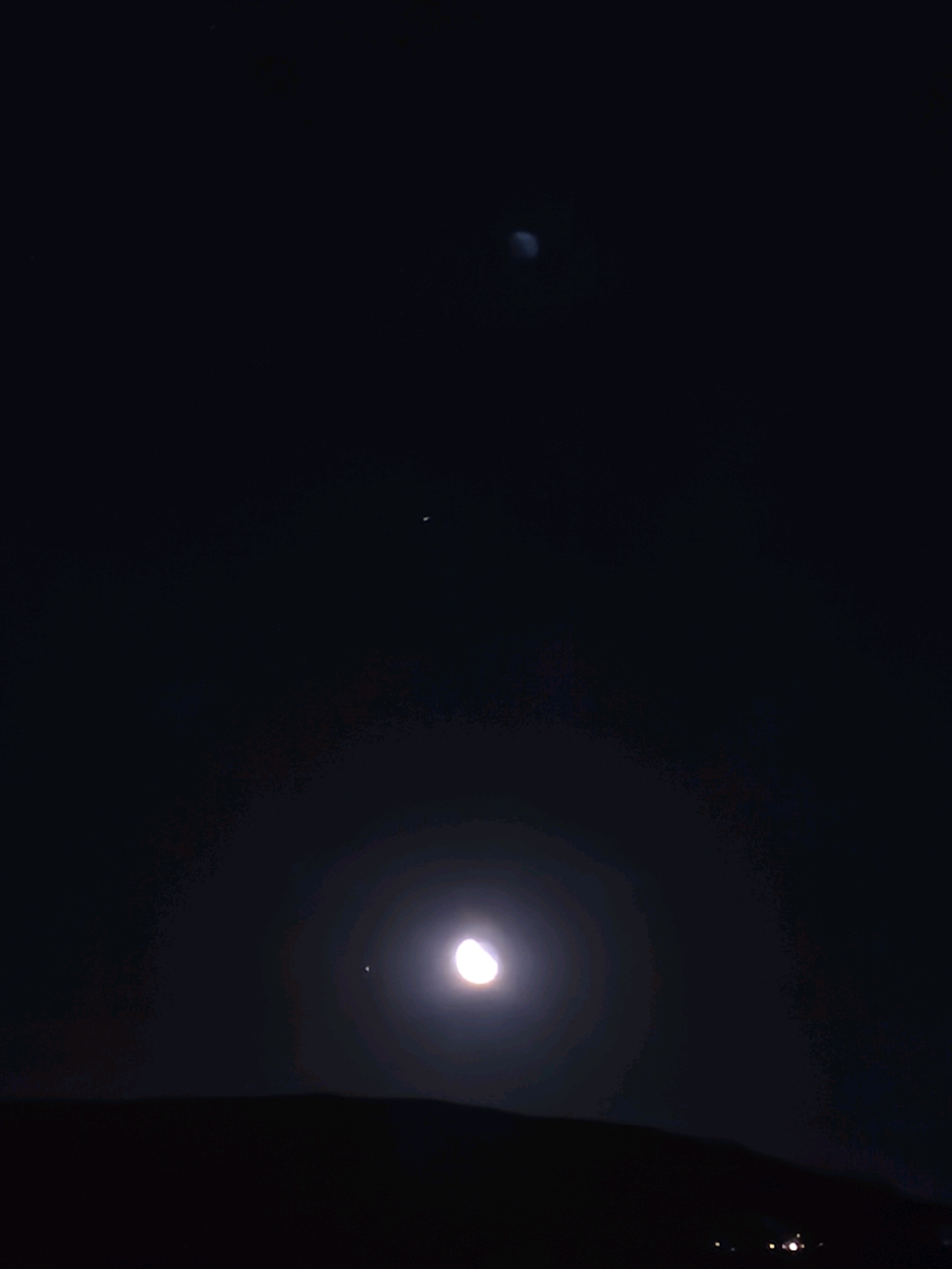 This is a nighttime image taken from Passignano sul Trasimeno, Italy, showing a view of the night sky over a dark, hilly landscape. In the foreground, a dark silhouette of hills is visible. The sky is dark with celestial bodies. At the bottom center of the frame, a bright crescent moon is prominent, surrounded by a luminous halo. To the left of the moon, a small, bright point of light, possibly a planet or star, is visible. Further up in the sky, near the top center, another small, faint point of light is present. Towards the top right of the frame, a larger, blurry oval shape, possibly another celestial object or an artifact of the photograph, is visible. The scene appears tranquil and captures a moment of astronomical observation.
