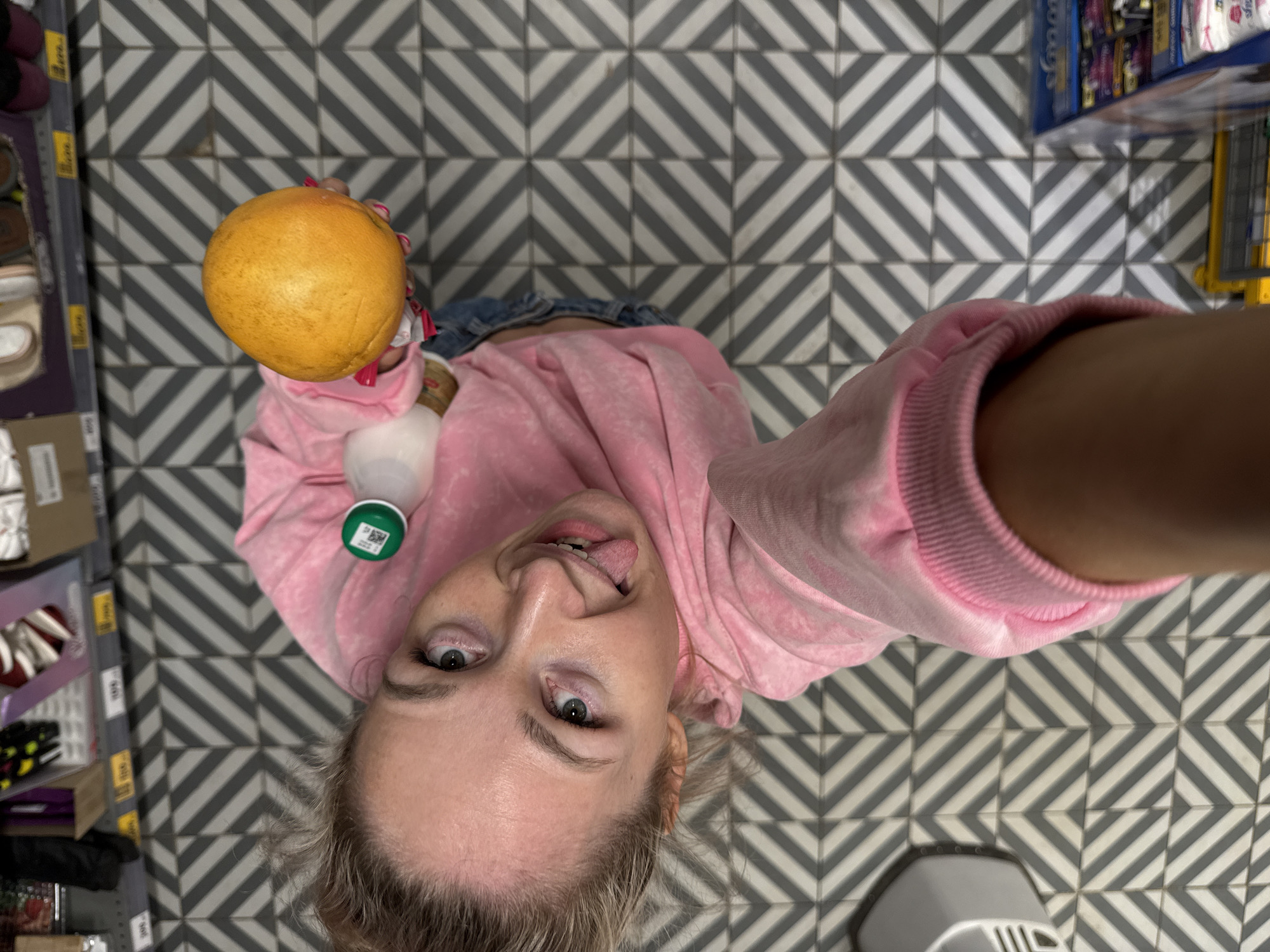 A selfie taken from an overhead perspective shows a fair-skinned woman holding a spherical orange fruit and a bottle of milk. She is wearing a light pink hooded sweatshirt. The woman's face is visible, her eyes are open, and she is sticking out her tongue. The background is a tiled floor with a zigzag pattern of white and gray tiles. Partial views of store shelves are visible on the left and right sides of the frame, suggesting the setting is a supermarket aisle. A shopping cart is visible at the bottom. This image was taken in the city of Chaikovsky, Russia.