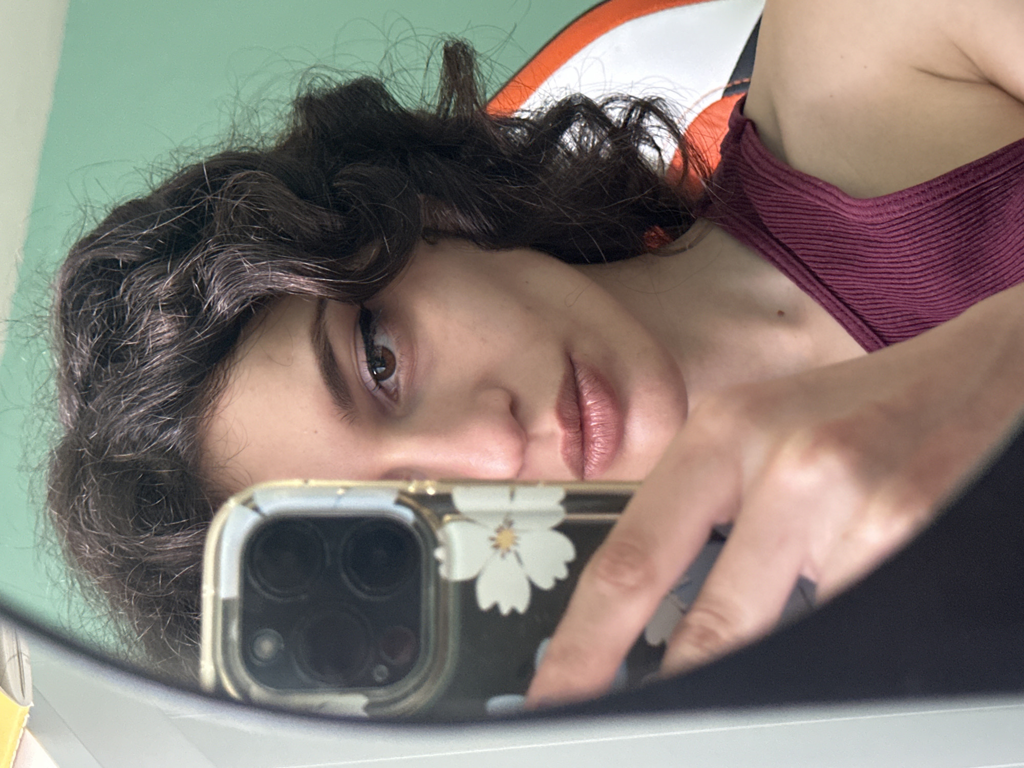 A medium shot shows a person's face reflected in a mirror as they take a selfie. The person has dark, curly hair and is wearing a dark pink, ribbed tank top. They are looking directly at the camera with makeup on their eyes and lips. The background is a light green wall. The image is taken in Sofia, Bulgaria.