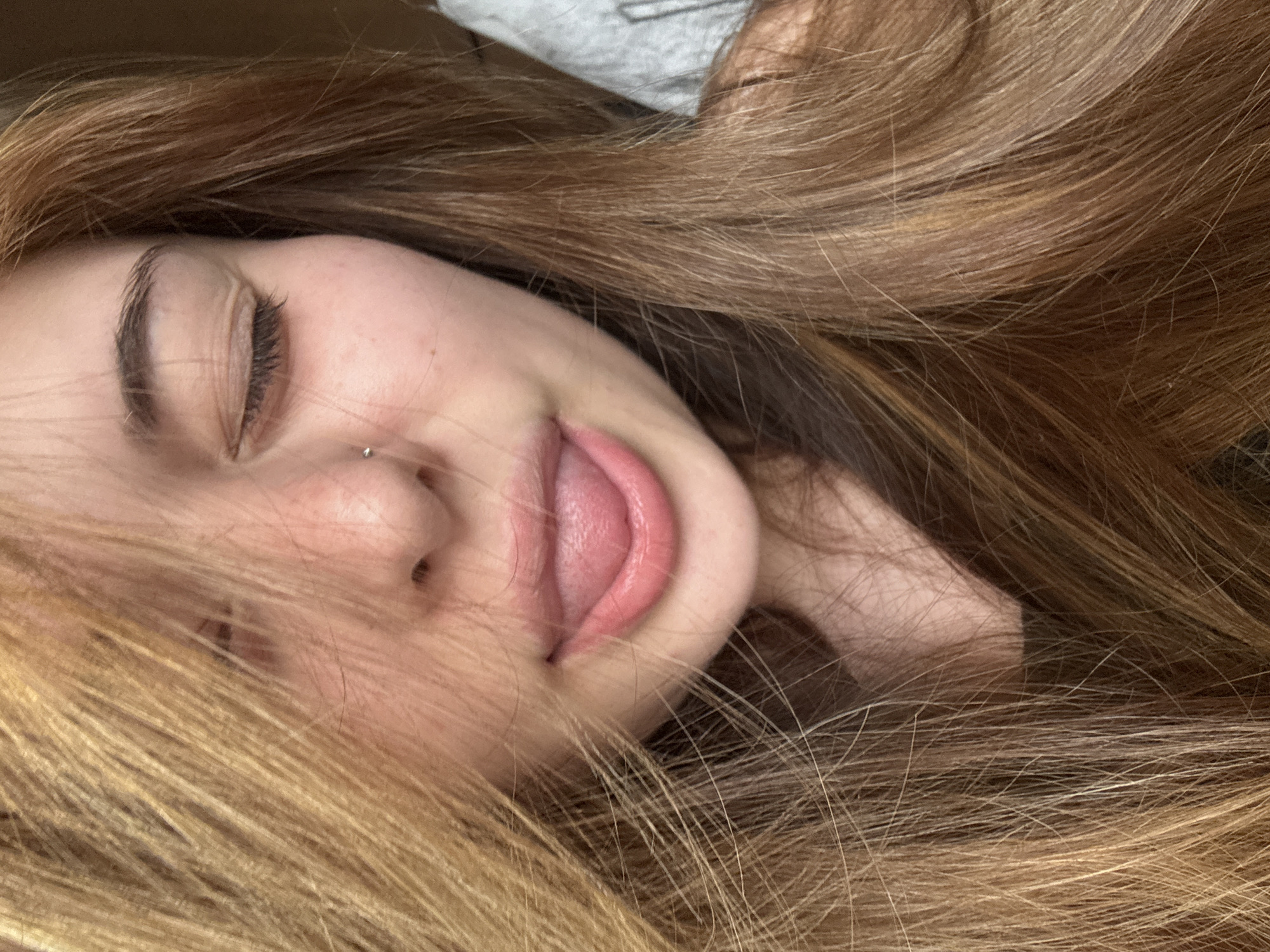 A close-up portrait captures a young person, likely female, lying down in what appears to be a relaxed or playful pose. Her face is partially obscured by her long, wavy, light brown to auburn hair, which is fanned out around her head. Her eyes are closed, emphasizing her dark, well-defined eyebrows and long eyelashes. A small, metallic nose piercing is visible on her right nostril. Her lips are slightly parted, revealing a portion of her tongue, suggesting a casual, comfortable, or perhaps whimsical expression. Her skin is fair, with faint freckles on her nose and cheek area. In the upper left corner, a sliver of light grey, textured fabric, possibly part of clothing or bedding, can be seen, hinting at an indoor setting. The lighting is soft and diffused, casting gentle shadows and creating an intimate atmosphere. No discernible text is visible in the image. While the location context of Troyan, Bulgaria is provided, there are no specific visual cues within the image itself to indicate this geographical location. The overall impression is one of tranquility, peace, or comfortable repose.