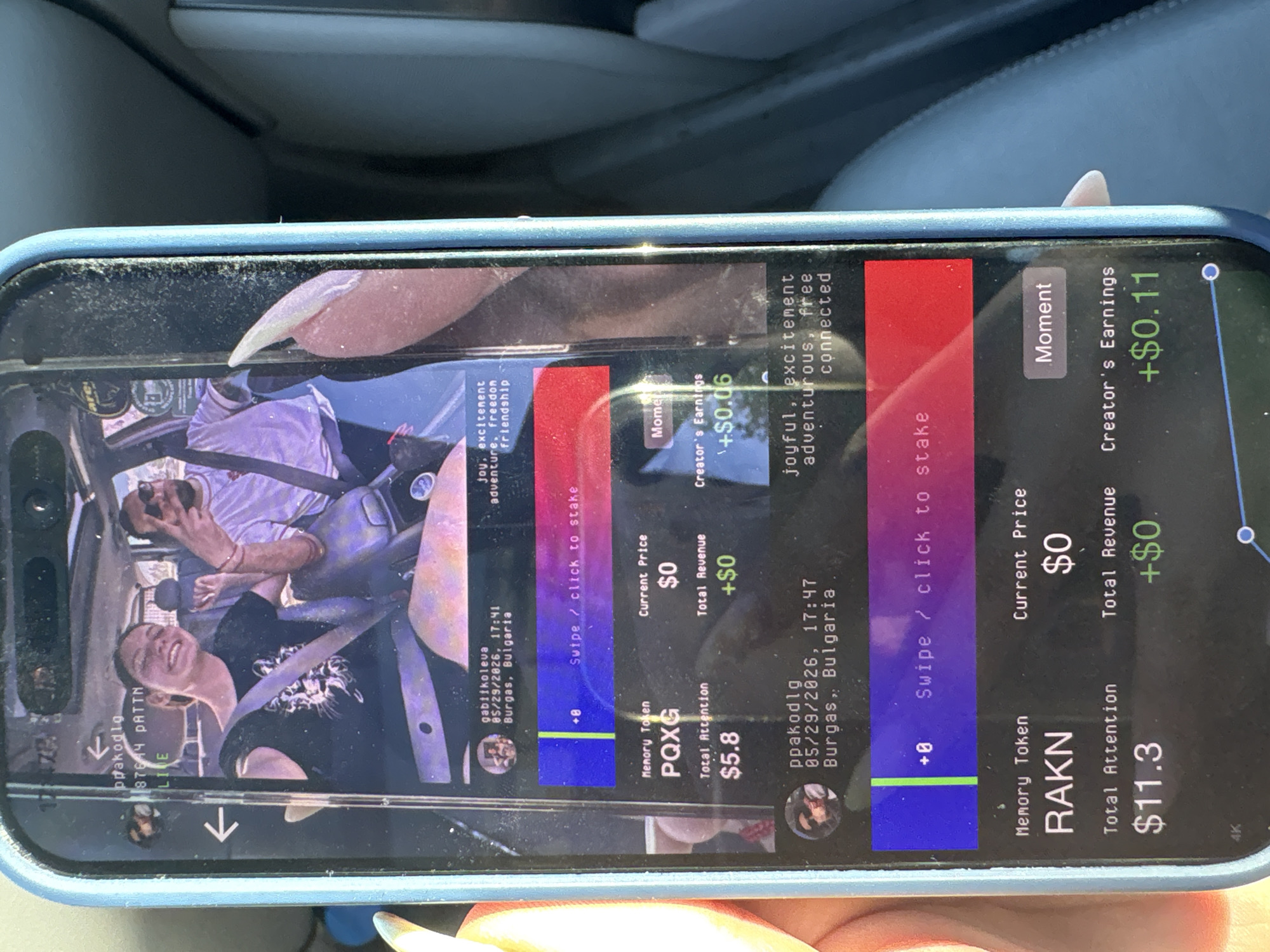 The image shows a smartphone screen displaying a social media or content platform. The location "Burgas, Bulgaria" is mentioned in the context of the content creators "gabiikoleva" and "ppakodlg" and the date 05/29/2026.
On the screen, the content creators are shown using a dashboard with memory tokens. gabiikoleva’s memory token is "PQXG" with a total attention of $5.8, a current price of $0, and a total revenue of +$0. ppakodlg's memory token is "RAKN," with a total attention of $11.3, a current price of $0, and a total revenue of +$0. In a separate part of the dashboard, there are creators’ earnings of +$0.06 for one user and +$0.11 for the other. The interface includes calls to action like "Swipe/click to stake."
An image of two people in a car is displayed on the screen.