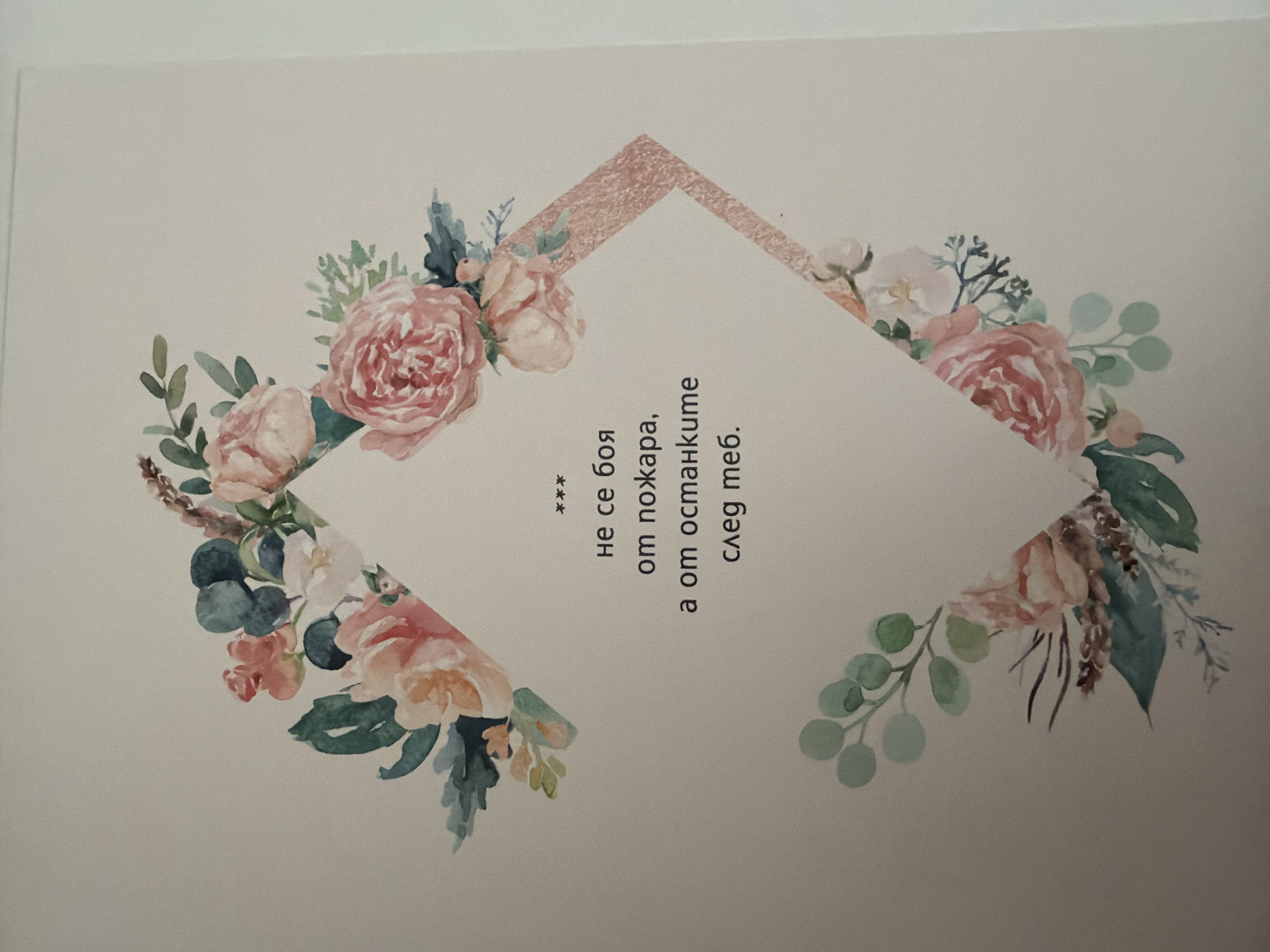 The image displays a card with a floral watercolor design and text in Bulgarian. The design features a symmetrical arrangement of pink and white roses, eucalyptus leaves, and other foliage, framing a diamond-shaped space in the center. The text, positioned within this central space, reads: "*** не се боя от пожара, а от останките след теб." This translates to: "*** I am not afraid of fire, but of the remains after you." The background of the card is a plain, light-colored surface, likely white or off-white. There are no people or specific locations in Pazardzhik, Bulgaria depicted in the image.