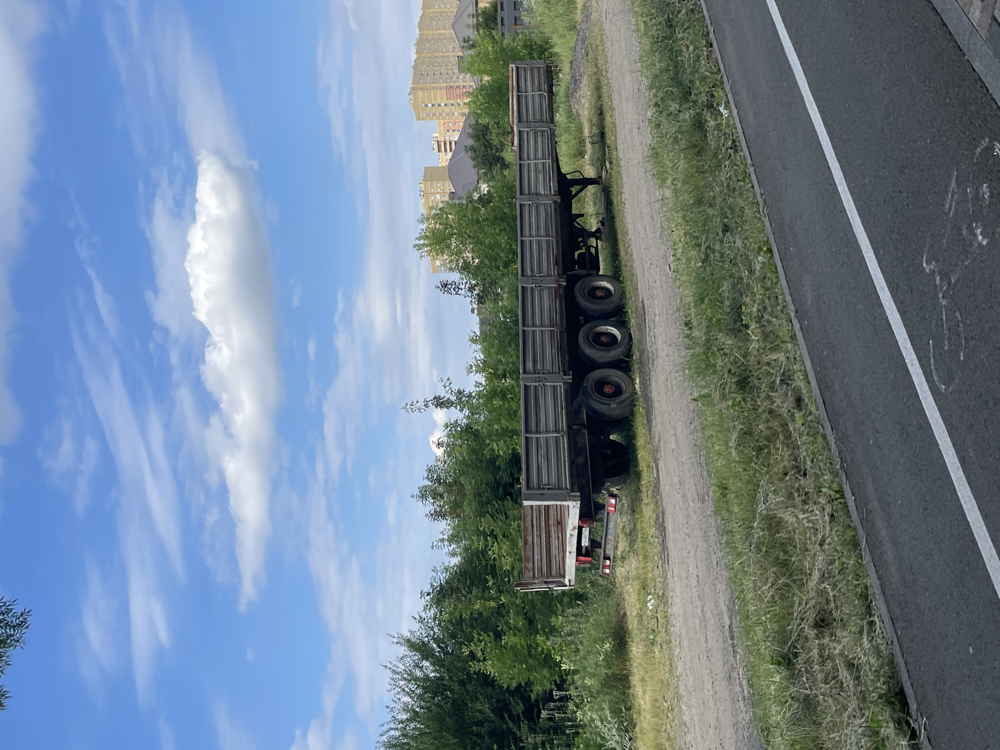 The image shows a parked, empty flatbed trailer with six wheels, positioned on a dirt and grass area adjacent to a paved road. The trailer has a metal bed with high sides and appears to be in a state of disrepair, with some rust visible on its surfaces. Behind the trailer, a dense line of green trees and foliage stretches across the mid-ground. In the background, several multi-story residential buildings, light yellow and beige in color, rise above the trees under a blue sky with scattered white clouds. The scene is outdoors, with a focus on the trailer and its surroundings. No people or animals are visible. The lighting suggests it is daytime, likely during the summer months due to the lush green vegetation. Based on the architectural style of the buildings and the overall appearance of the landscape, this scene could be located in the city of Stavropol, Russia.