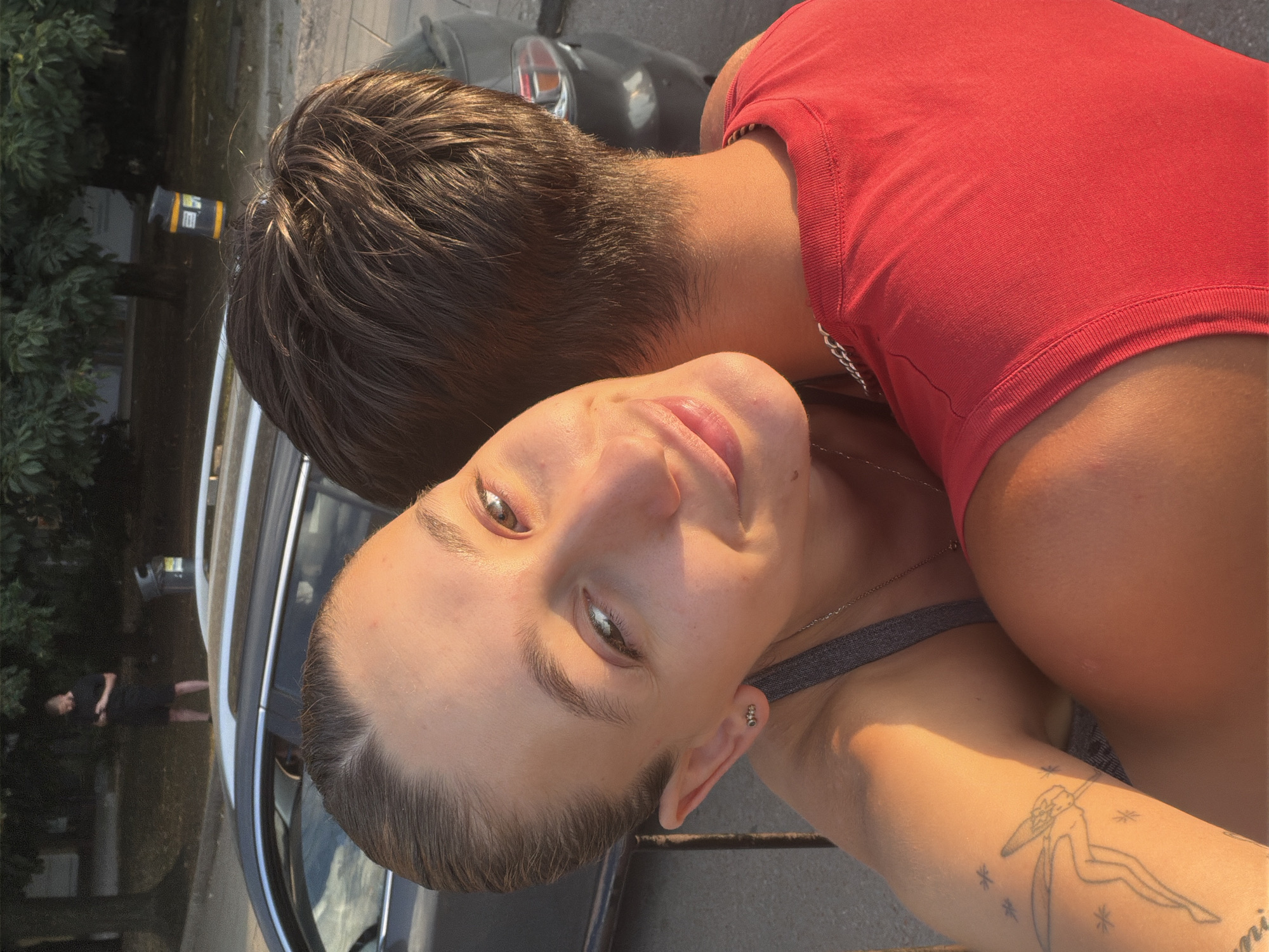 In Sofia, Bulgaria, a young woman with light skin and dark brown hair tied back, wearing a gray tank top and stud earrings, embraces a young man from behind. The man, whose face is obscured, wears a red tank top. Sunlight illuminates the scene. In the background, a dark-colored car is parked, and further back, a man in black shorts and a black t-shirt stands near a tree, looking at his phone. A yellow and black trash bin is also visible.