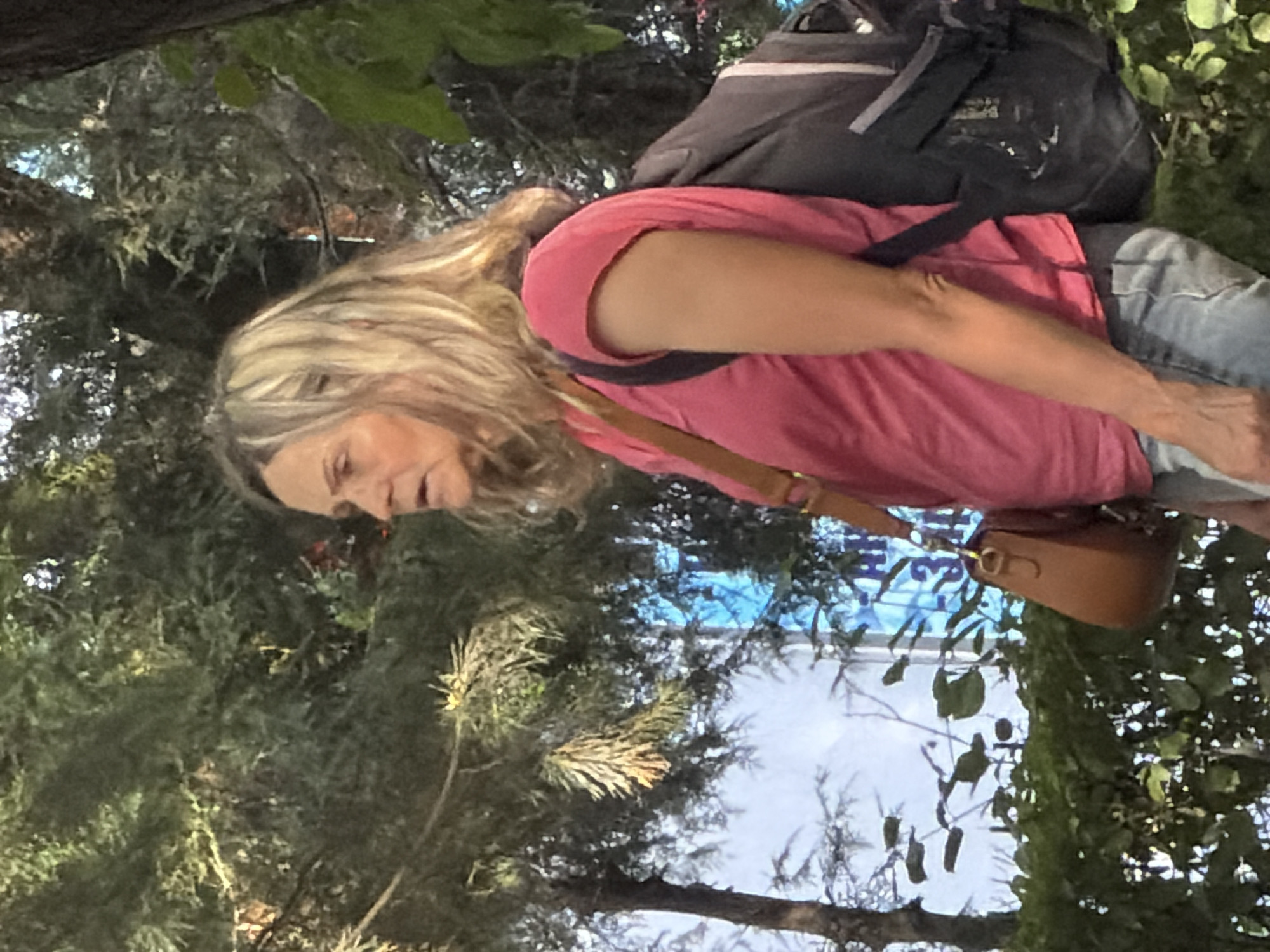 A medium shot captures a light-skinned woman in a pink t-shirt and blue jeans, seen from the chest up, walking outdoors. Her blonde hair is shoulder-length and styled with loose waves. She wears a brown crossbody bag and a dark backpack. The background consists of blurred green foliage and trees, with a glimpse of a blue sign with white text. The scene appears to be an outdoor urban environment.