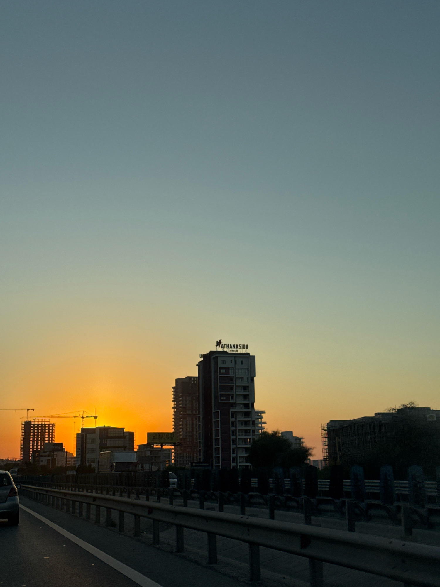 The image captures a sunset over a city skyline, likely in Mouttagiaka Community, Cyprus. The sky transitions from a pale blue at the top to a vibrant orange near the horizon where the sun is setting. In the foreground, a highway is visible with a metal guardrail. On the right side of the highway, there are trees and various buildings, including a tall building with "ATHANASIOU TOWER" signage. On the left side of the highway, multiple construction sites are present with cranes and unfinished buildings. A silver car is partially visible on the left edge of the frame, traveling on the highway. The scene appears to be a dusk setting with an urban landscape.