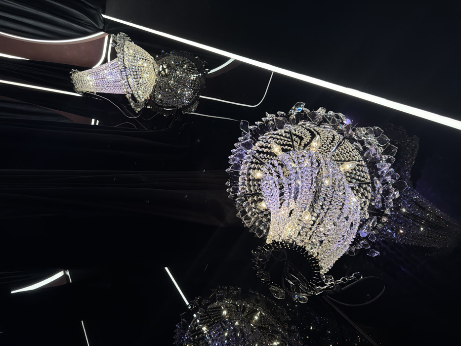 This image captures a dramatic indoor scene featuring multiple ornate crystal chandeliers against a deep black, highly reflective background. The composition highlights the sparkling brilliance of the chandeliers and their sharp reflections.
In the upper left quadrant, two smaller, bell-shaped crystal chandeliers are visible, one appearing brighter and more iridescent, the other darker with a glittery effect. These are reflected in the dark surface below. The dominant feature is a large, spherical crystal chandelier occupying the right side of the frame. This chandelier is intricately designed with countless faceted crystals and small light bulbs emitting a warm, golden glow, causing bright highlights and prismatic effects across its surface.
The background is a smooth, glossy black surface that perfectly mirrors the chandeliers and linear light strips. These linear light sources, appearing as bright white lines, run across the top, middle, and bottom of the frame, also casting distinct reflections. The interplay of the bright, sparkling lights and the dark, reflective surface creates a sense of depth and luxury.
No people or specific activities are depicted, allowing the focus to remain solely on the decorative lighting and its visual impact. The setting evokes a sense of opulence and modern grandeur, characteristic of high-end venues. The artificial lighting suggests an interior space, likely a luxury store, exhibition, or architectural feature, aligning with the location context of Dubai, United Arab Emirates. The image conveys a sophisticated and dazzling atmosphere. No text is visible.