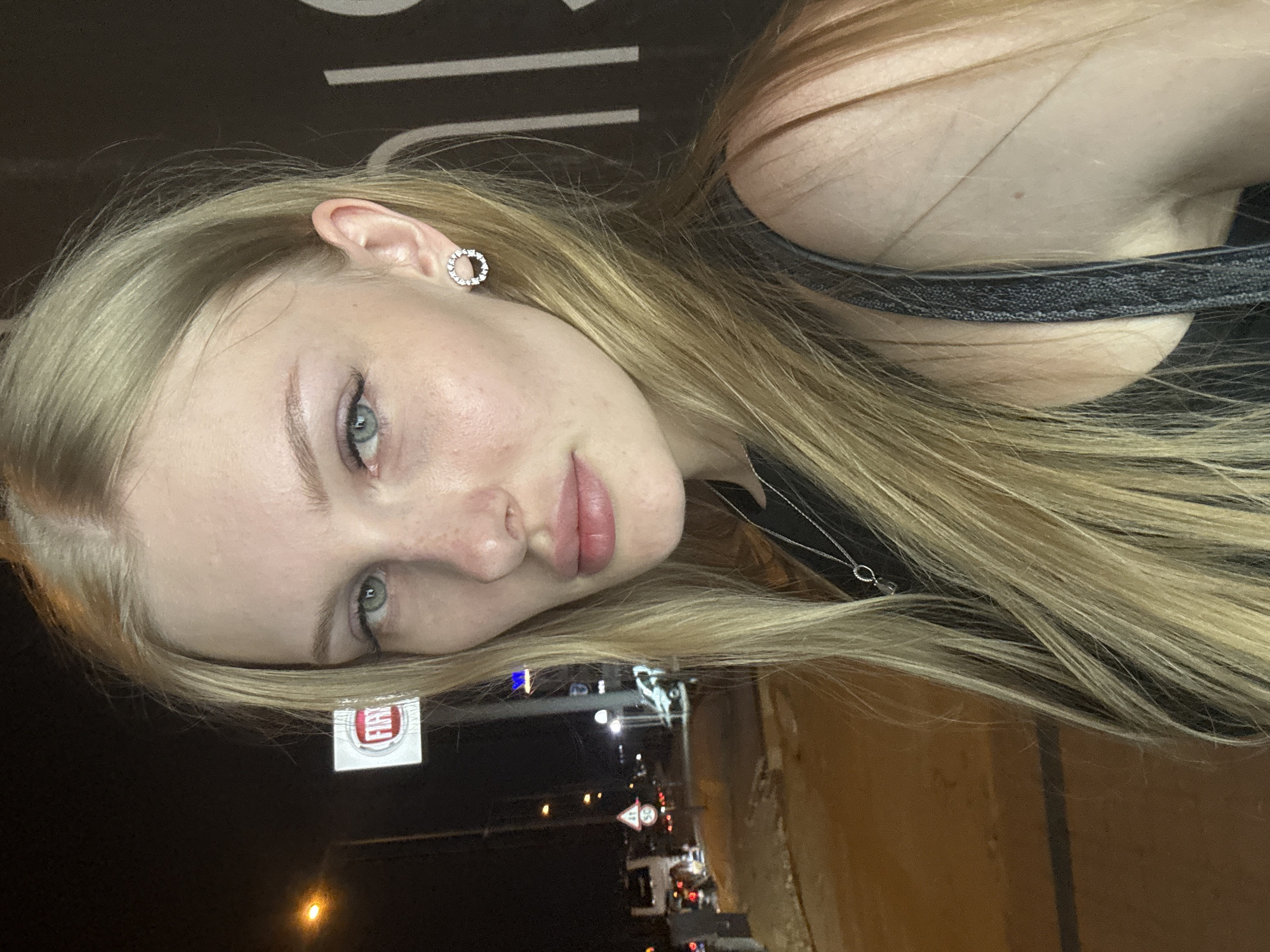 The image shows a close-up, front-facing selfie of a young woman with blonde hair and light blue eyes. She is wearing a black top and a silver necklace. Her right ear is adorned with a circular earring featuring small, clear stones. The background is dark with some light sources and signs, suggesting an outdoor urban environment at night. A portion of a red and white "FIAT" logo is visible on the left side, and a street with a few vehicles and traffic signs is discernible in the background, consistent with a location in Kepez, Turkey. The overall lighting is dim, with the woman's face being the primary illuminated subject.