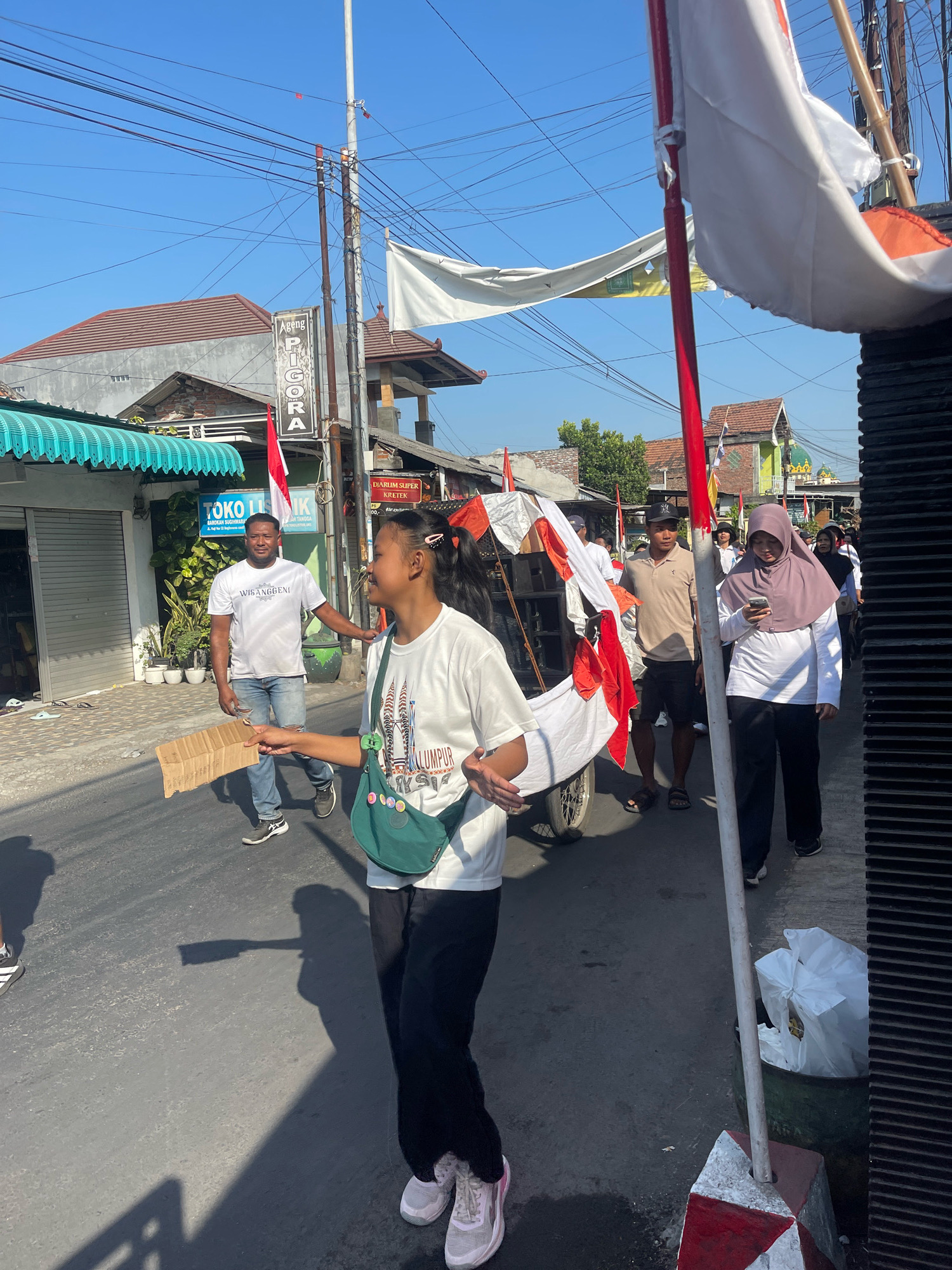 The image depicts a street scene in Candi, Indonesia, under a clear blue sky. In the foreground, a young woman with dark hair pulled back in a ponytail is walking, holding a piece of cardboard in her right hand. She is wearing a white t-shirt with graphics, a green crossbody bag adorned with circular pins, and black pants. Her left hand is gesturing outwards as she looks to her right. Behind her, a man in a white t-shirt and distressed blue jeans walks, looking towards the camera. To the right of the foreground subjects, a woman in a light purple hijab and white long-sleeved shirt is looking at her phone. Other individuals are visible in the background, including a man in a beige polo shirt and black shorts. A cart decorated with red and white flags is present, along with several Indonesian flags. Shops and buildings line the street, featuring signage that includes "Ageng PIGORA" and "TOKO LISTRIK." Overhead, a network of power lines crisscrosses the sky, and trees are visible in the distance. The street itself is paved, and shadows are cast on the asphalt.