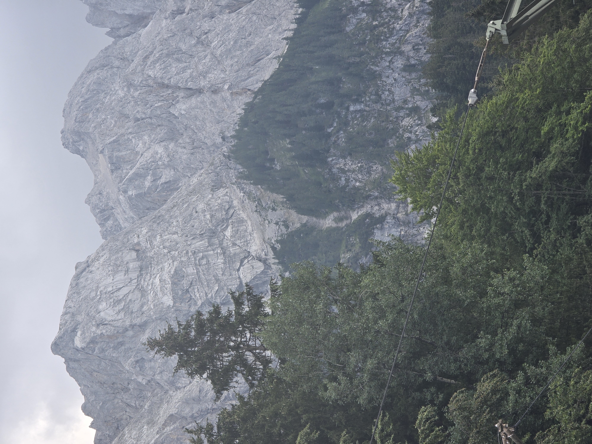 This is a vertical, medium shot of a mountain range with trees in the foreground. The mountains are rocky and gray, with some patches of snow or ice. They are partially obscured by a thick layer of clouds, which appear to be white and gray. The trees in the foreground are green and lush, with a variety of deciduous and coniferous trees. There is a cable extending across the lower part of the image, which is part of a cable car system. The image appears to have been taken in the Alps, possibly near Grainau, Germany, known for its cable car access to Zugspitze, Germany's highest peak. The scene is natural and scenic, with no people or animals visible.