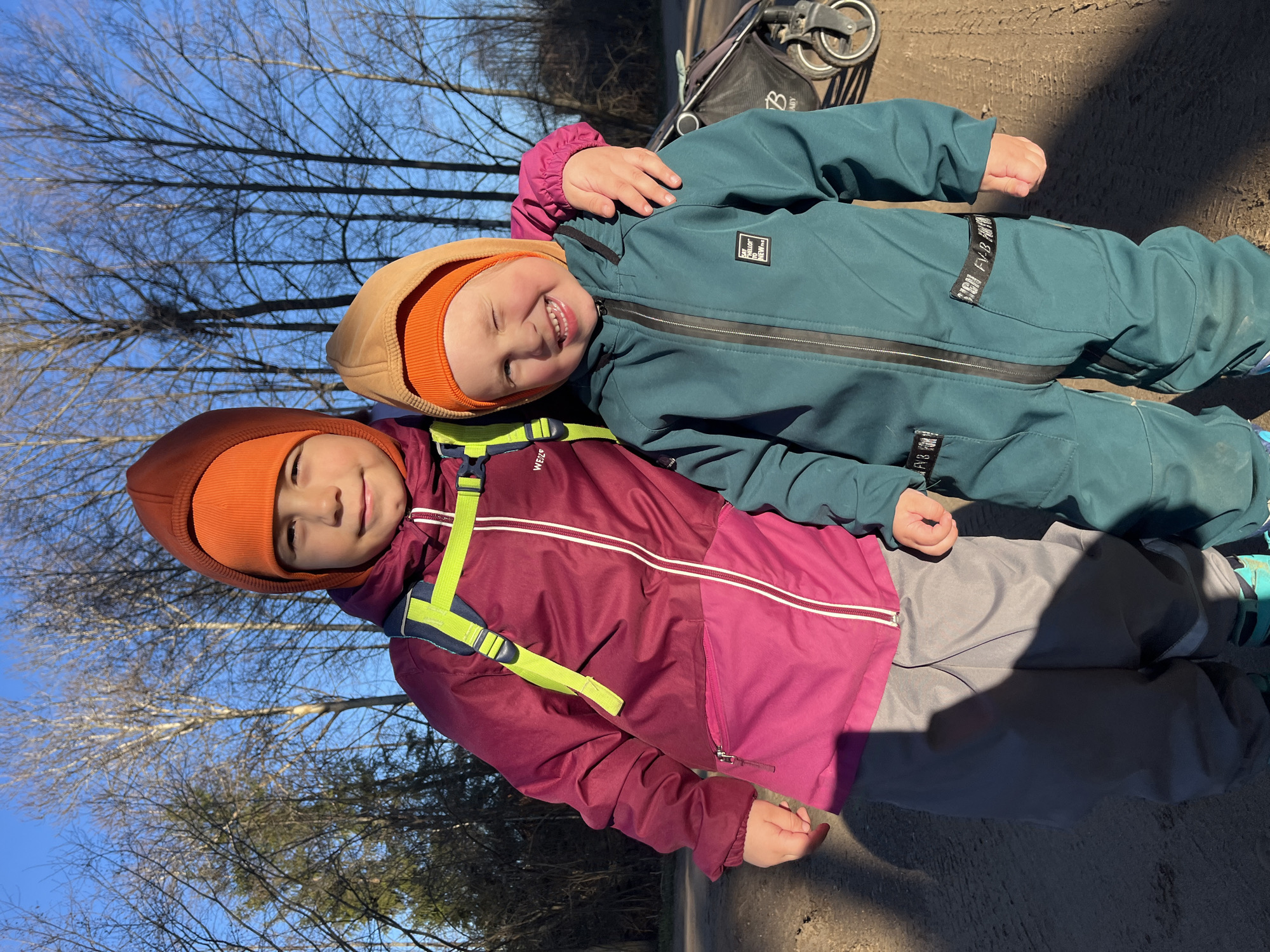 Two young children are captured in an outdoor setting on a sunny day in Chaikovsky, Russia. Both children are smiling broadly, suggesting happiness and playfulness. The child on the left wears an orange beanie under a maroon and pink hooded jacket, with a neon green strap from a backpack or harness partially visible, possibly from the brand "Wed'ze." The child on the right has their arm draped over the other's shoulder and is wearing an orange beanie under a light brown hood, along with a teal-colored full-body snowsuit or overall. This child is winking or squinting, likely due to the bright sunlight, and displays a wide, joyful grin.
The background features numerous bare tree branches against a clear, bright blue sky, indicating late autumn, winter, or early spring. The ground appears to be dry dirt or gravel, with long shadows cast by the strong sunlight. A black stroller or buggy, featuring a "B" logo, is partially visible on the right side of the frame. Visible text on the right child's jacket includes "SAY HELLO NEW" on a white patch, and "FVB" repeated on a black strap around their leg. The overall impression is a cheerful moment shared between the children during an outdoor activity on a clear, cold day.