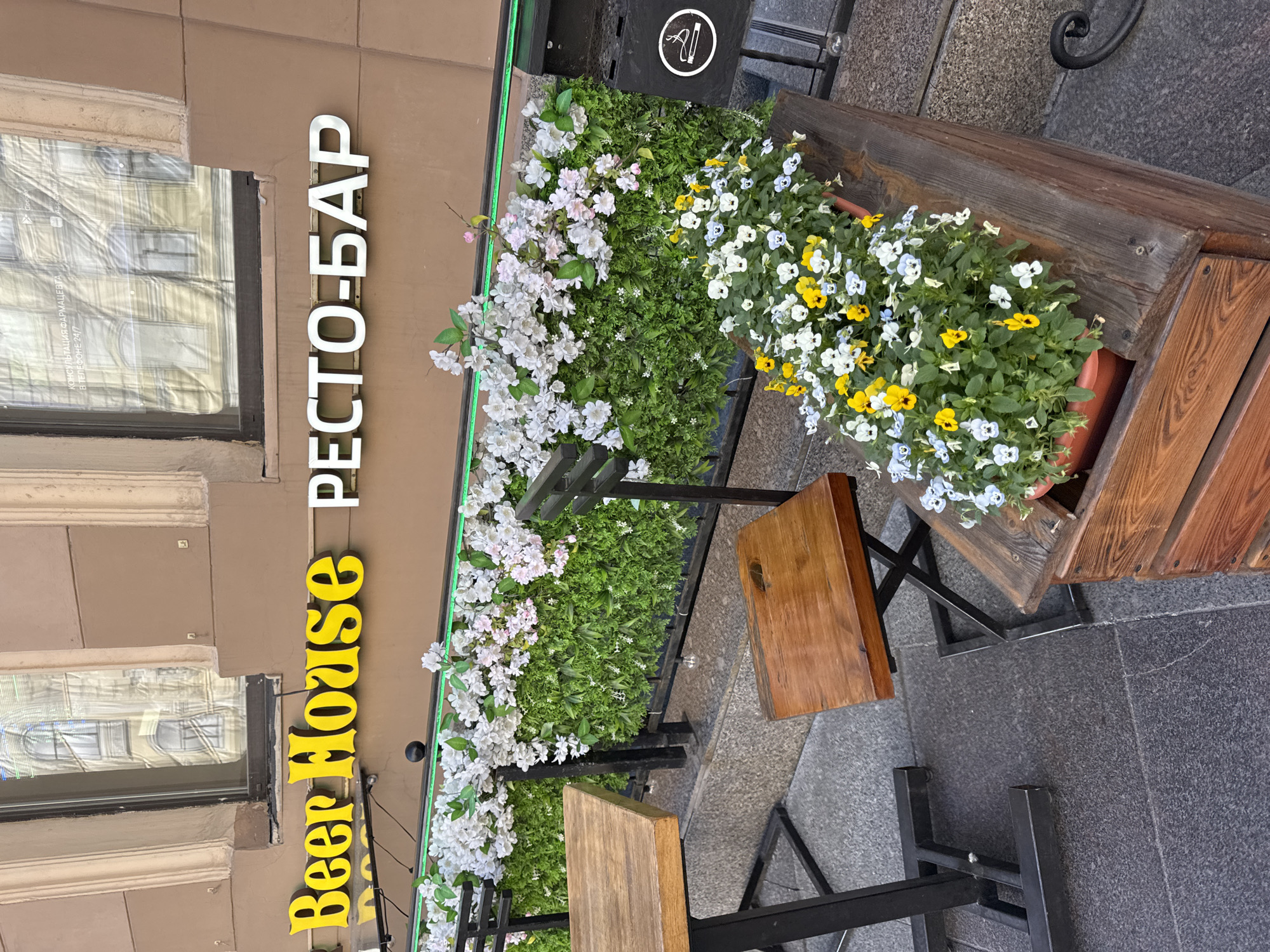 The image shows the exterior of a establishment named "Beer House РЕСТО-БАР" in Saint Petersburg, Russia. The sign for "Beer House" is in yellow serif font, and "РЕСТО-БАР" is in white sans-serif font. The establishment has an outdoor seating area with a table and chairs made of wood and metal. There are planters with green foliage and white and pink flowers, as well as a planter with white, yellow, and blue pansies. A black ashtray with a no-smoking symbol is visible on the right side of the image. The building facade is light brown with windows reflecting surrounding buildings. The ground is paved with granite tiles.