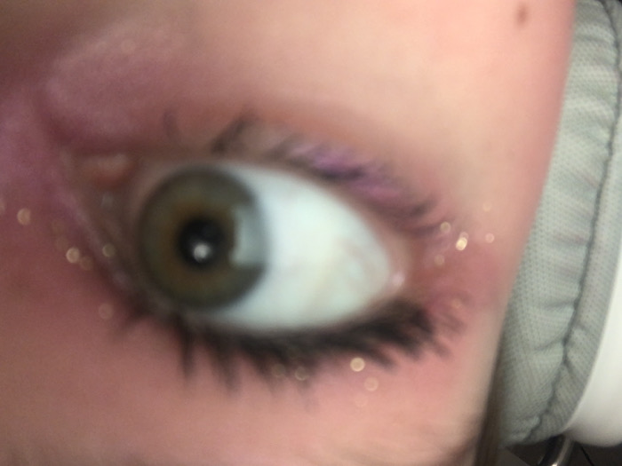 A close-up captures a hazel eye, partially blurred. The eye is rimmed with dark lashes, and traces of light purple eyeshadow are visible. A sprinkling of small, shiny particles, possibly glitter, adds sparkle around the eye. Pinkish skin surrounds the eye, and the edge of a gray, padded object, possibly headphones, appears on the right side of the frame.
