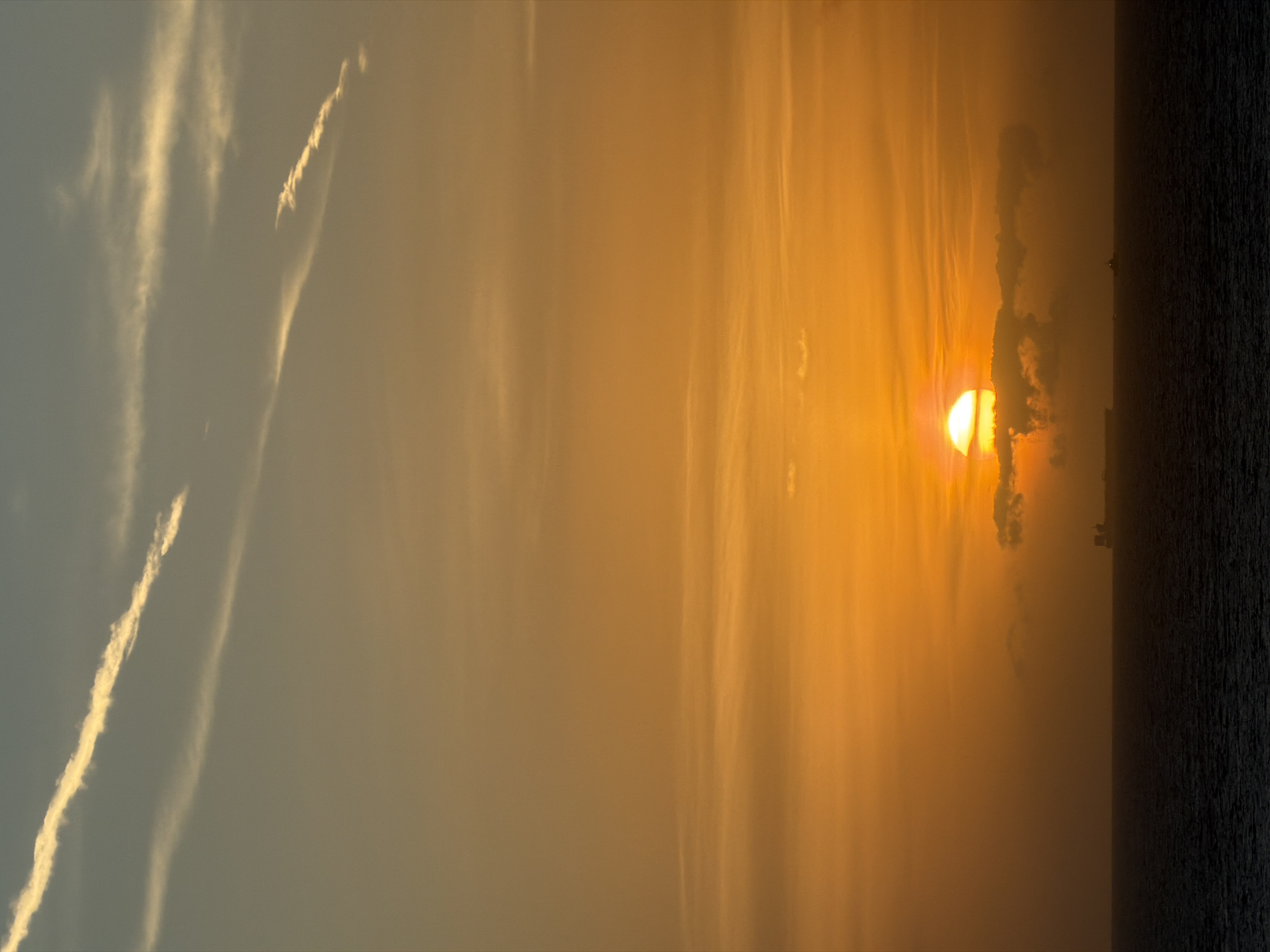 A vertical, daytime shot shows the sun as a bright orange orb through clouds. The sky transitions from grey at the left to orange where the sun shines. A dark area suggests water and land at the right. Streaks in the sky appear to be contrails from airplanes. The location is Unknown, Unknown.