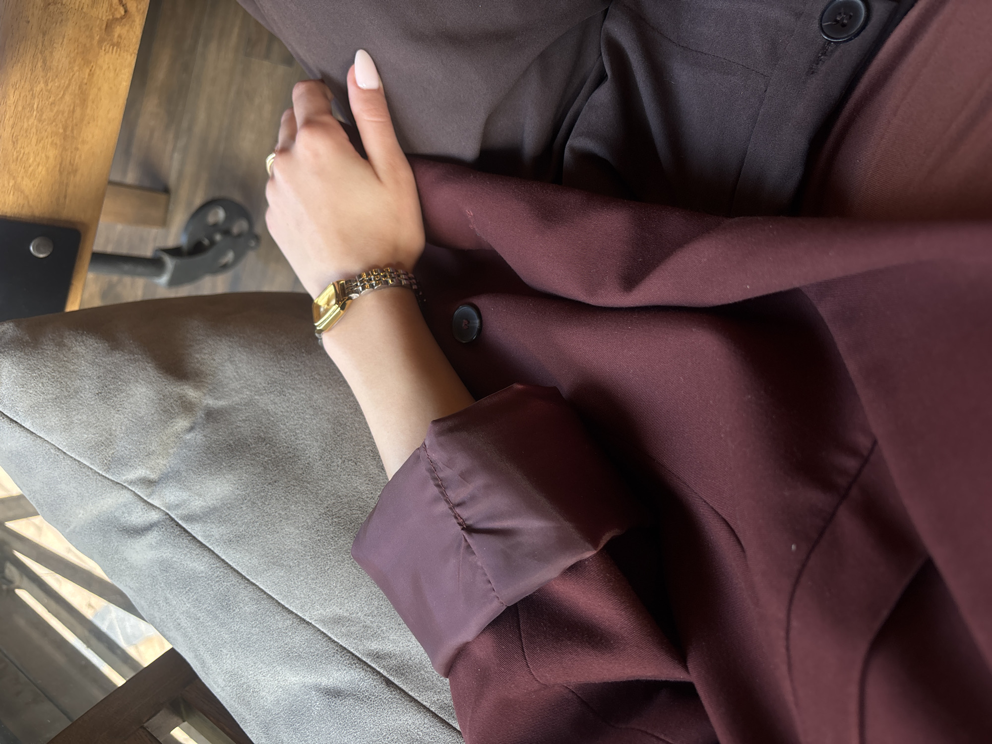 The image shows a close-up shot of a person's arm and clothing. The person is wearing a gold-colored wristwatch and a ring on their finger. They are wearing a burgundy-colored blazer over a brown shirt. The person's hand rests on the blazer, near the button. The background consists of a gray cushion and a wooden surface, possibly a table or chair. The location is unknown.