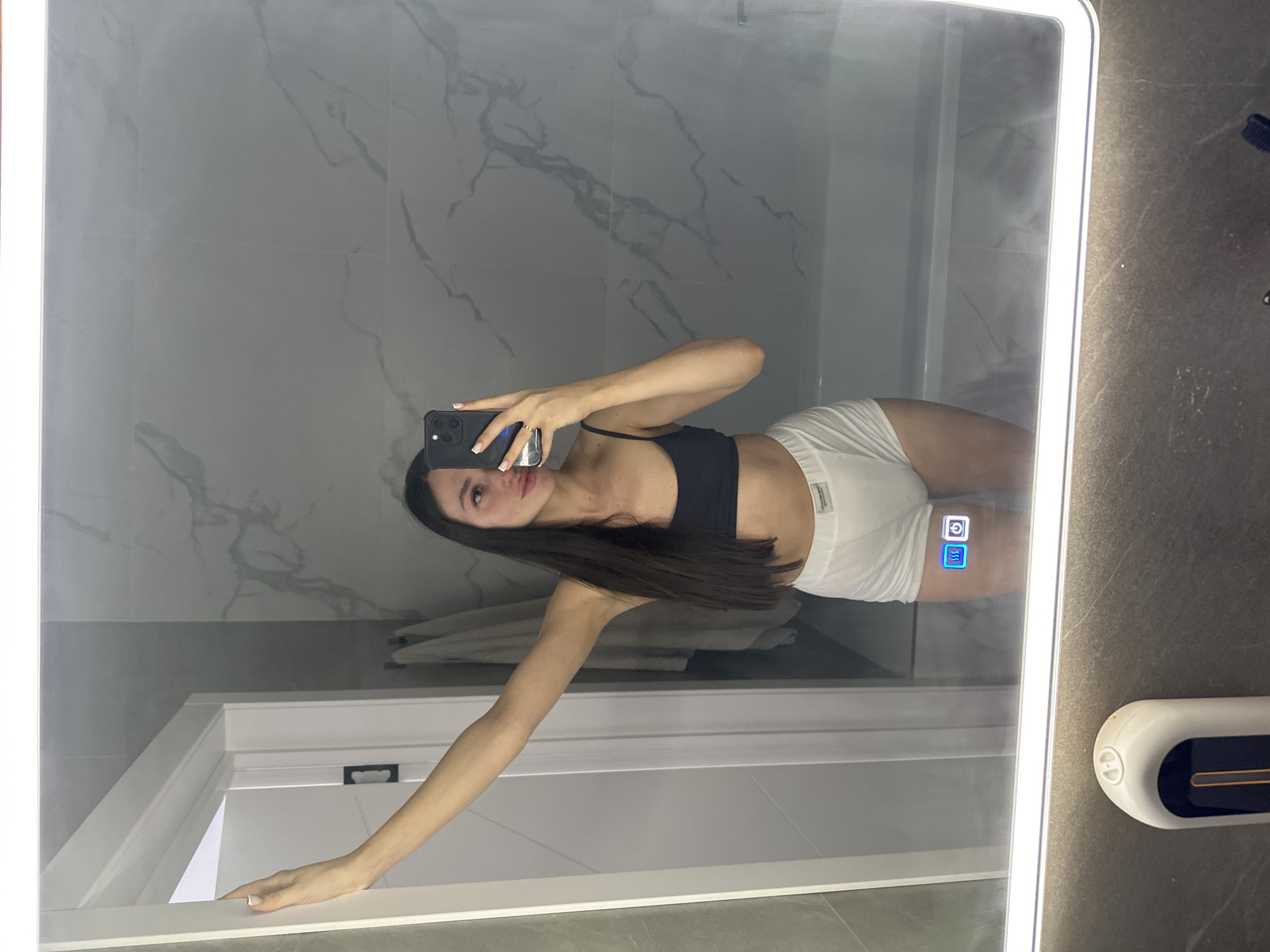 A full-length mirror selfie is taken by a woman with long, dark hair in a bathroom. She is wearing a black sports bra and white high-waisted shorts. She is standing with her left arm extended upwards, leaning against a white doorframe. Her right arm is bent, holding a black smartphone to take the photo. The background shows marble-patterned tiles on the wall and a white bathtub. The mirror has a built-in light around its frame and displays two illuminated icons near the bottom: a camera icon and a power icon. In the foreground, a portion of a white automatic soap dispenser is visible. The scene is in the city of Estachi, Russia.