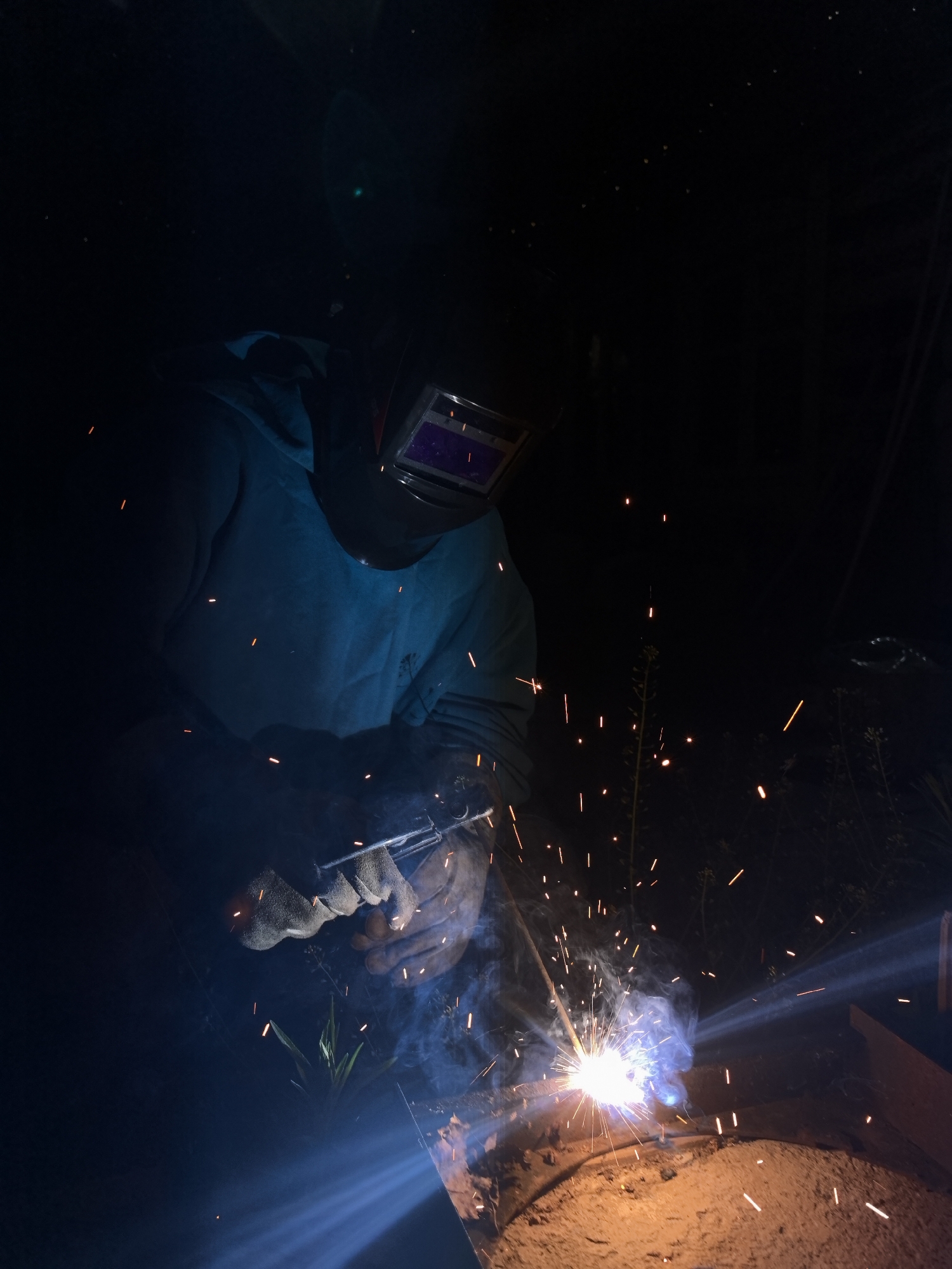 A welder in protective gear is working in the dark, with sparks and light emanating from the welding point. The location information is not discernible from the image.
