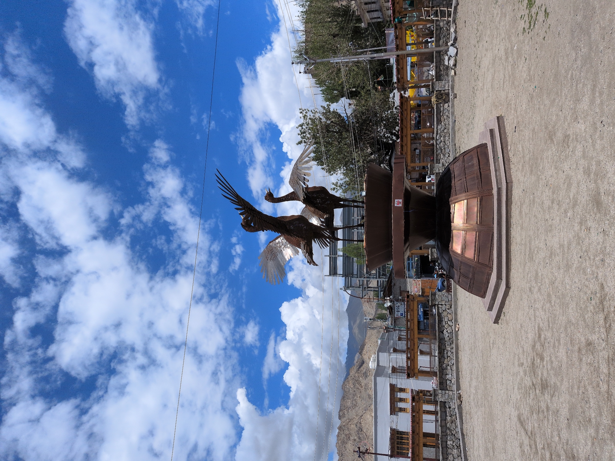 This image depicts a public square in the city of Leh, India, featuring a prominent bronze sculpture of two cranes in flight. The sculpture is centrally located on a tiered, octagonal base, also made of bronze, which is surrounded by a gravel-covered ground. In the background, traditional Ladakhi architecture is visible, characterized by flat roofs and wooden balconies. Mountains form the backdrop, suggesting the city's high-altitude location. The sky is a vibrant blue, dotted with fluffy white clouds. Utility poles and wires are present, indicating the urban setting. A few people are visible in the distance, engaged in activities typical of a public space. The overall impression is one of a well-maintained cultural landmark within a bustling city.