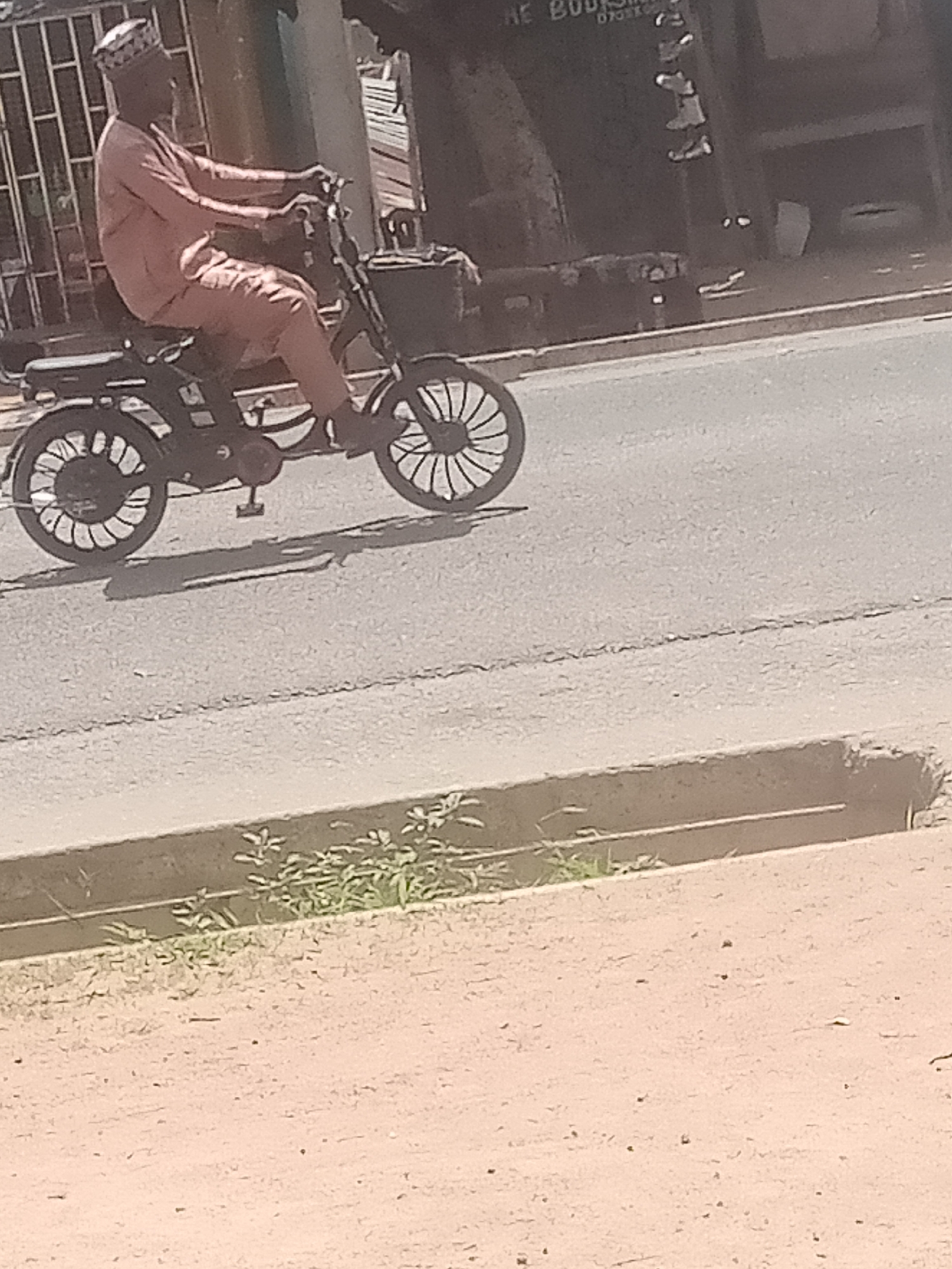 A man wearing a light pink traditional outfit and a patterned cap rides a dark-colored motorbike. He is seen from the side, looking to his left. The motorbike has distinctive spoked wheels. The scene appears to be an outdoor urban setting, possibly a street in Jalingo, Nigeria. The ground is paved, and there is a concrete curb or drainage channel to the right of the road, with some sparse green vegetation growing in it. In the background, there is a dark-colored vehicle or structure with some visible text that is partially obscured and unreadable. The lighting suggests it is daytime, with shadows cast on the road, indicating the sun is present. No specific emotions are evident from the man's posture.